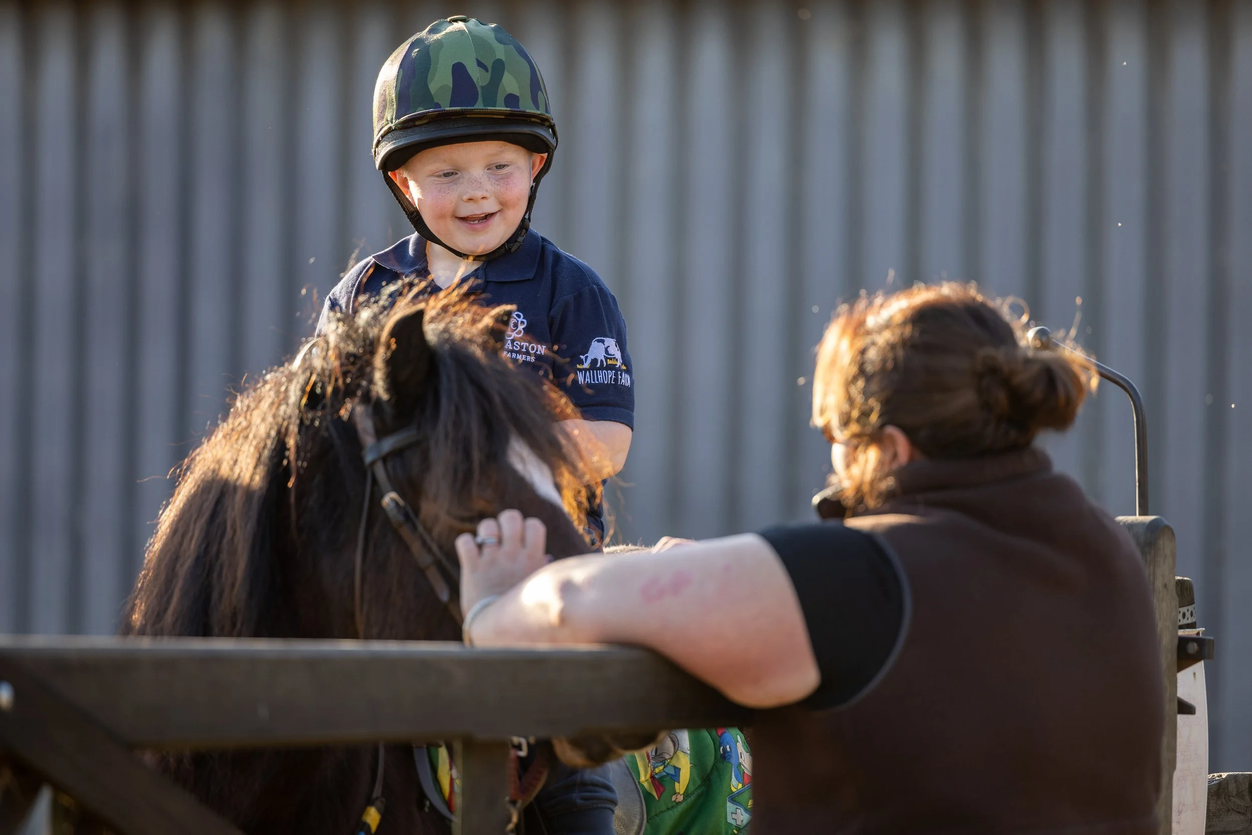 A young boy wearing a camouflage helmet riding a small pony, supervised by an adult woman with glasses, outdoors near a wooden fence.