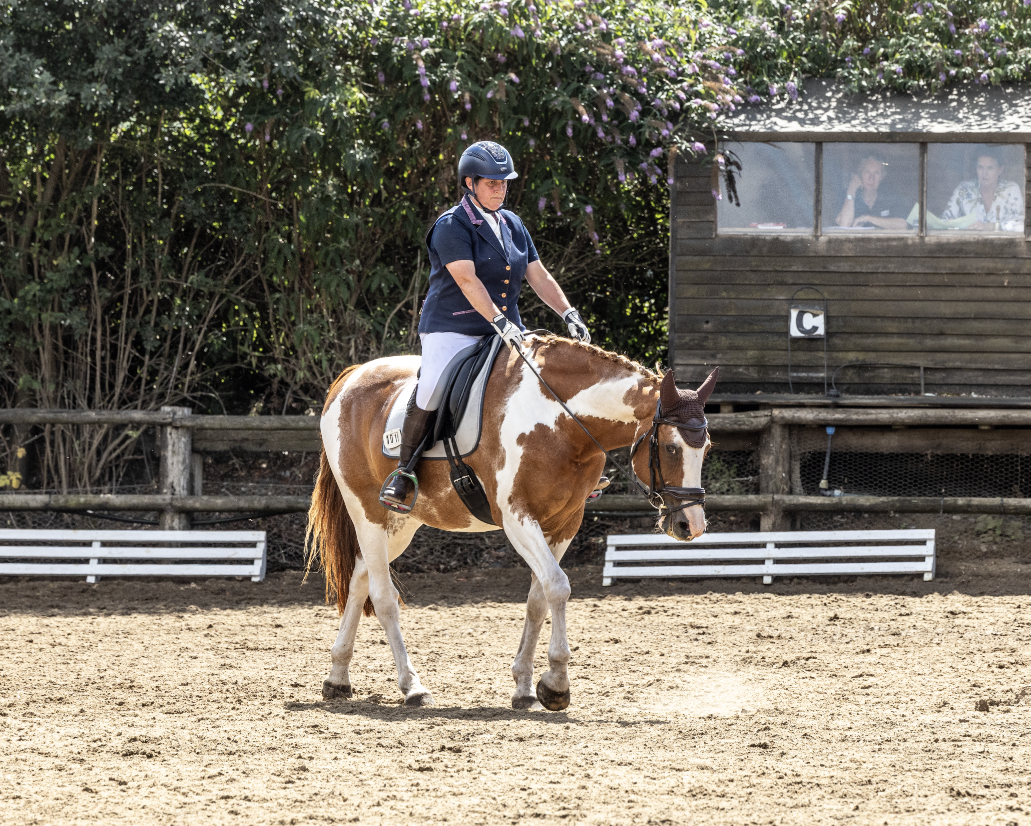 A woman riding a paint horse with a dark blue riding jacket, white riding pants, and a helmet, practicing in an outdoor riding arena. In the background, a wooden structure with windows shows two people seated inside.