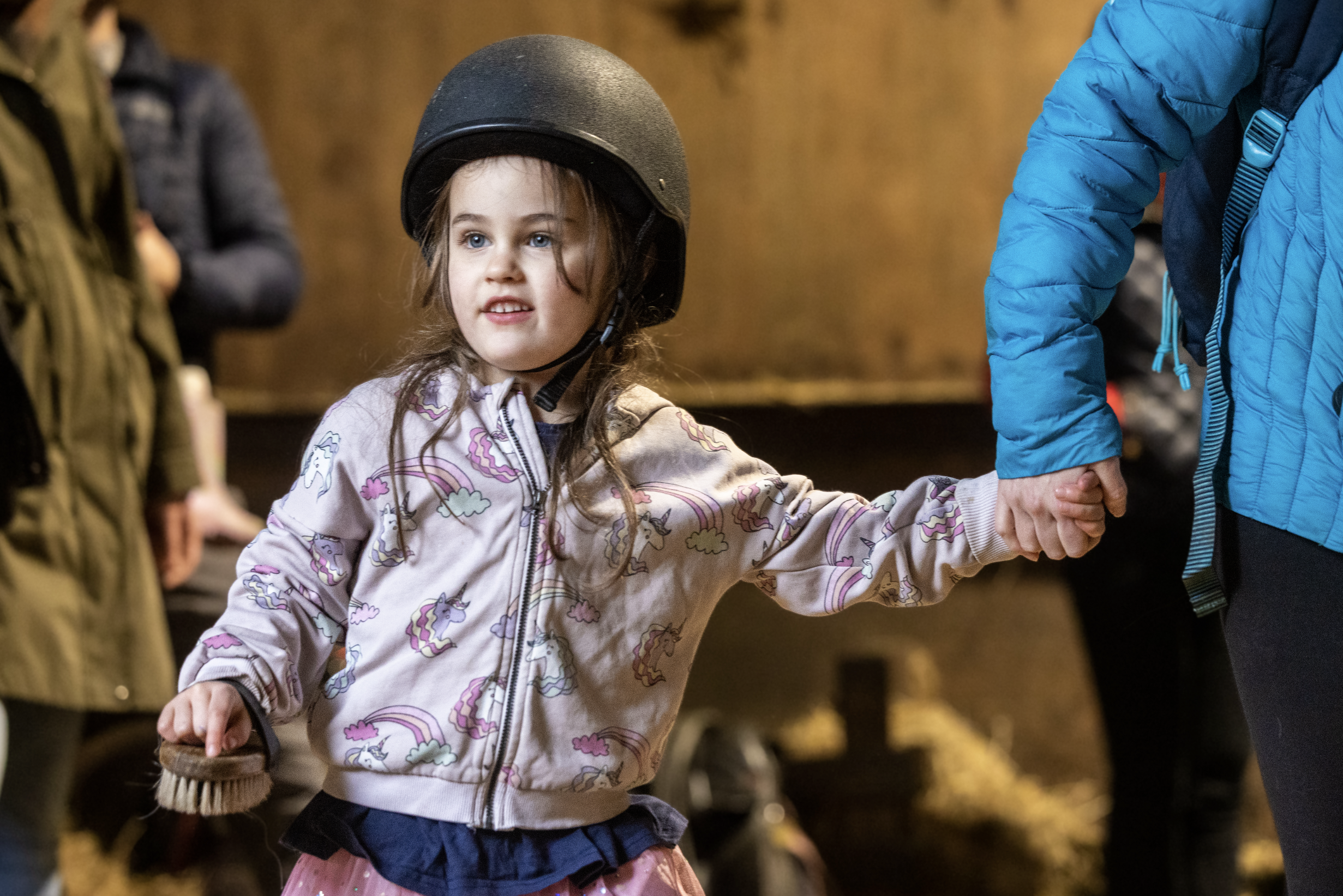 A young girl wearing a black helmet holding hands with an adult wearing a blue jacket, indoors with a sandy-colored wall and blurred background.