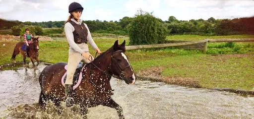 A girl riding a horse through a water crossing on a grassy field, with another rider in the background.
