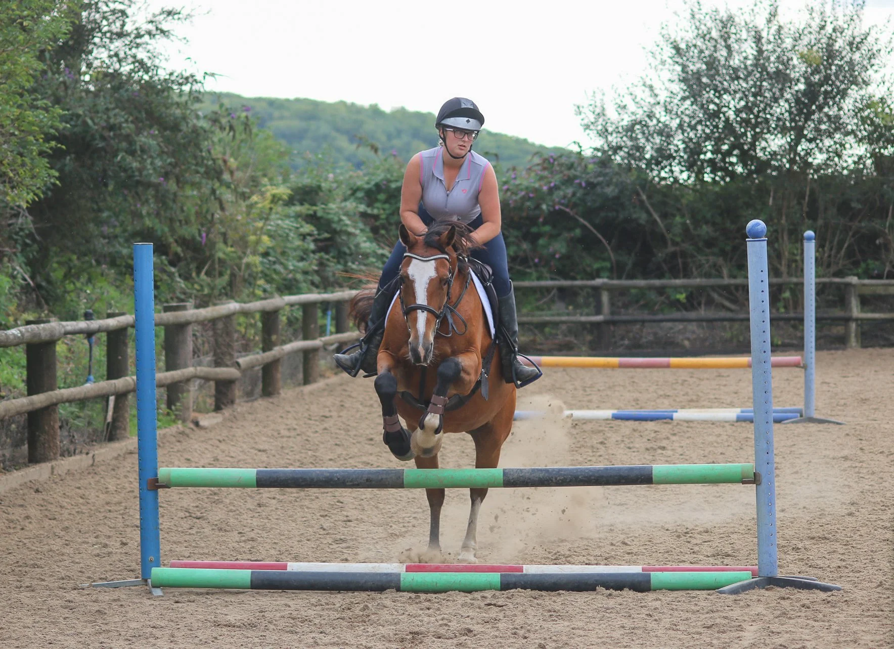 A woman riding a horse jumps over a colorful obstacle in an equestrian arena surrounded by trees and a wooden fence.