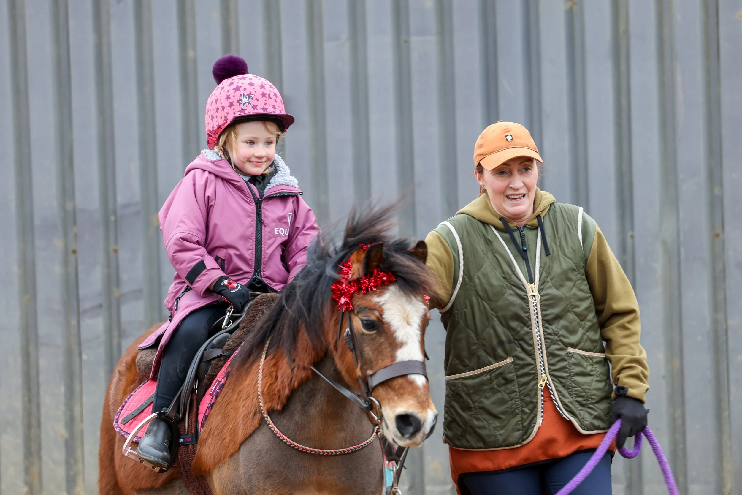 A young girl riding a brown horse with a woman walking beside them. The girl wears a pink jacket, pink hat with a pom-pom, and a helmet, smiling. The horse has red tinsel on its mane. The woman is dressed in a green vest, khaki hoodie, and orange cap