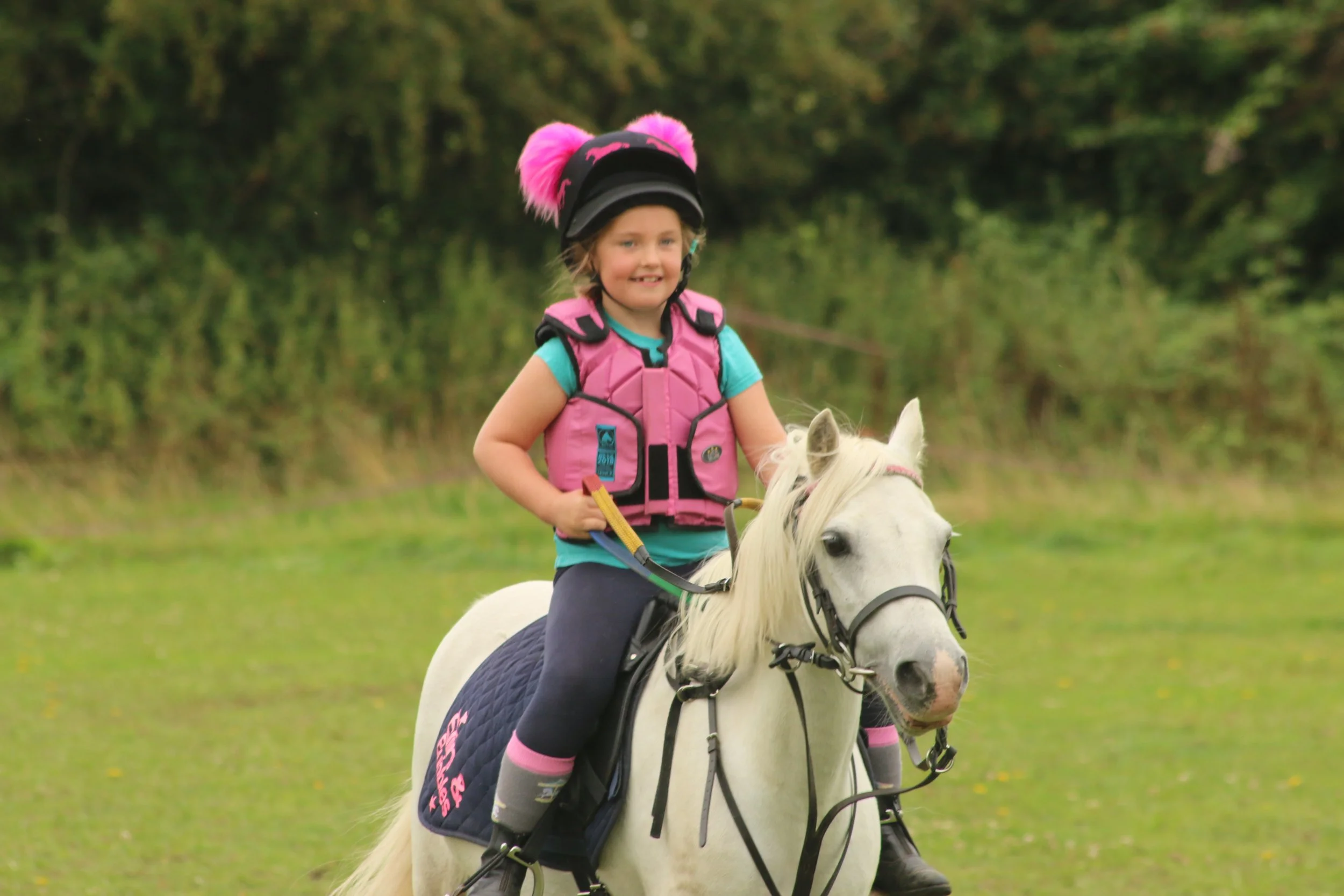 A young girl riding a white horse in an outdoor grassy area, wearing a pink helmet with fluffy pink ears and protective gear.
