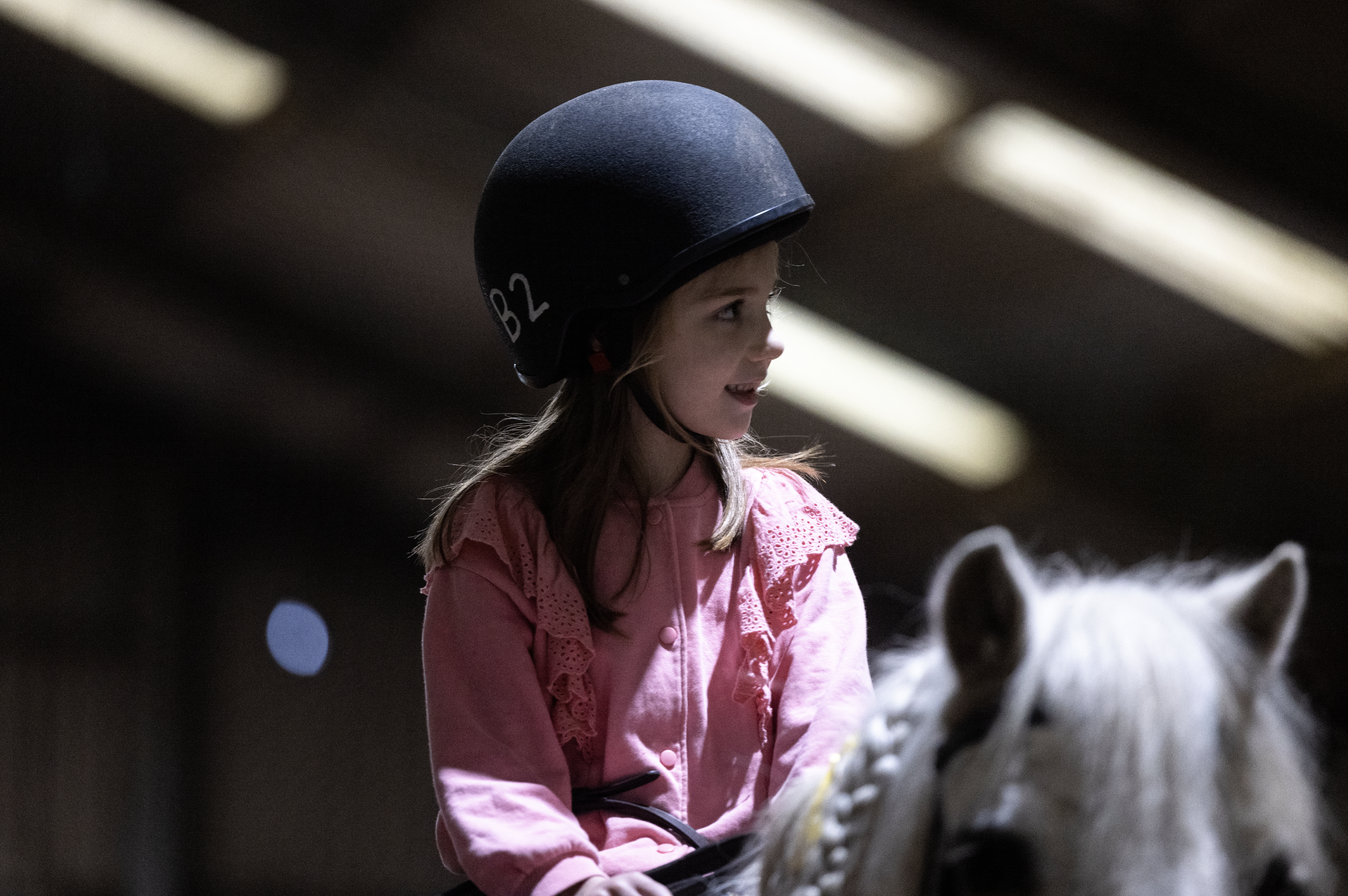A young girl wearing a black riding helmet and pink lace detail jacket, riding a white horse in an indoor riding arena.
