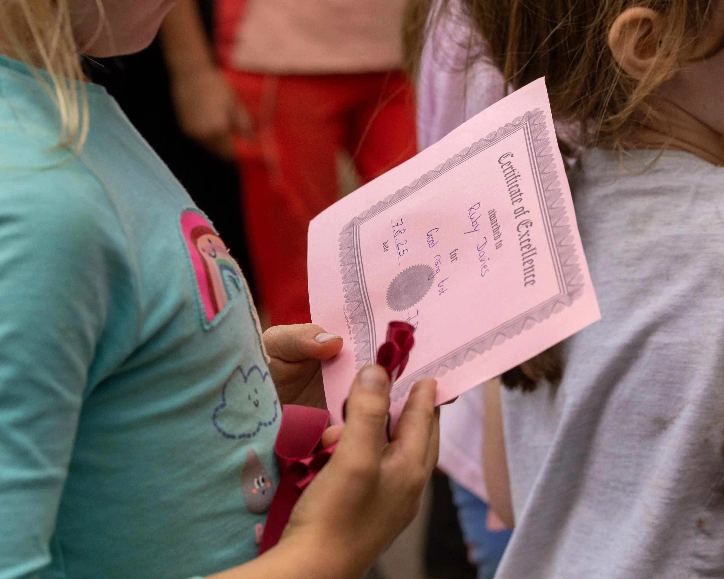 Child holding a pink certificate of excellence with a red ribbon, surrounded by other children.