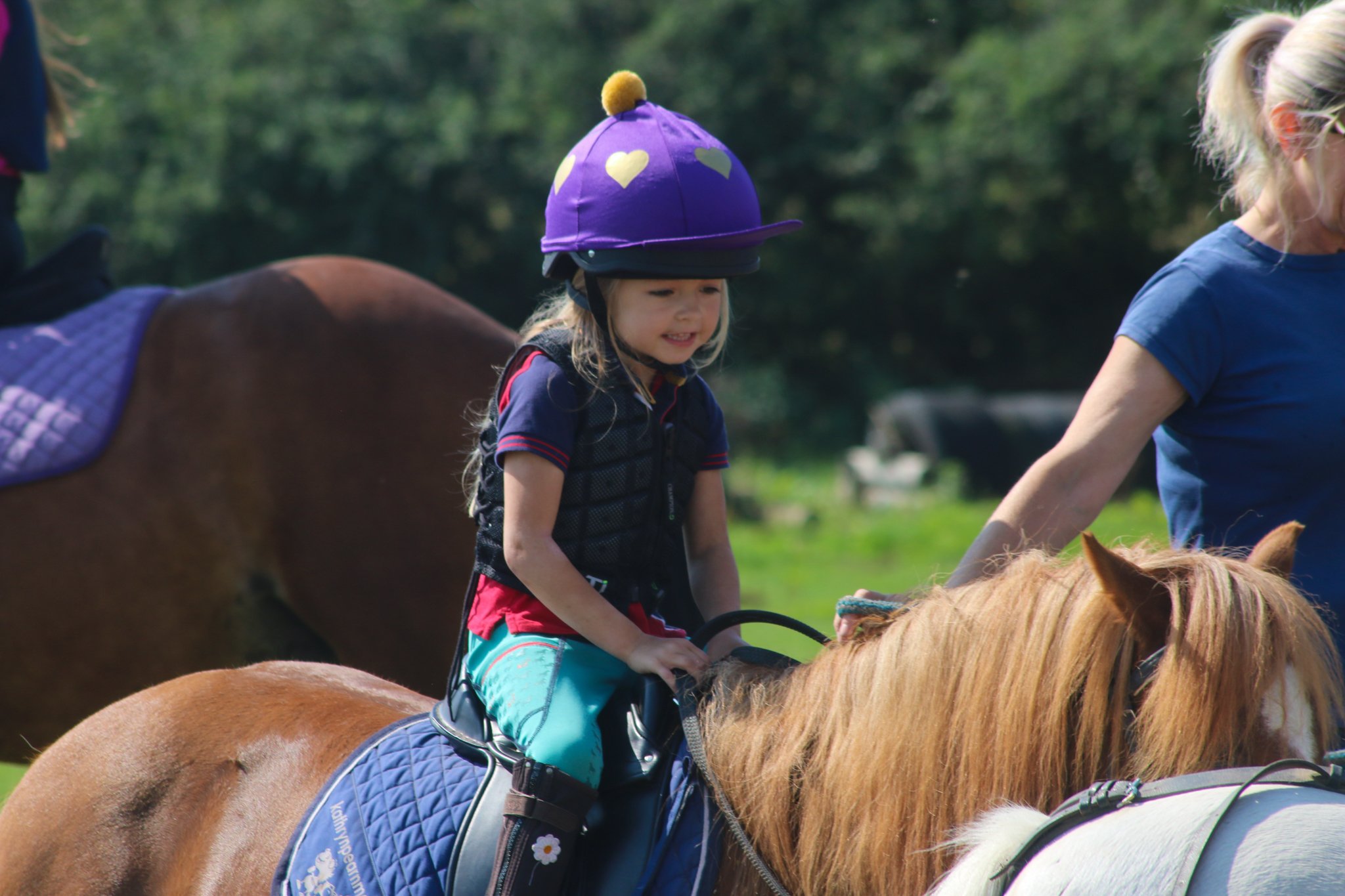 A young girl wearing a purple helmet with hearts rides a brown horse. An adult, partially visible, guides the horse, with just her arm and part of her face showing. The background is a green outdoor area with trees.