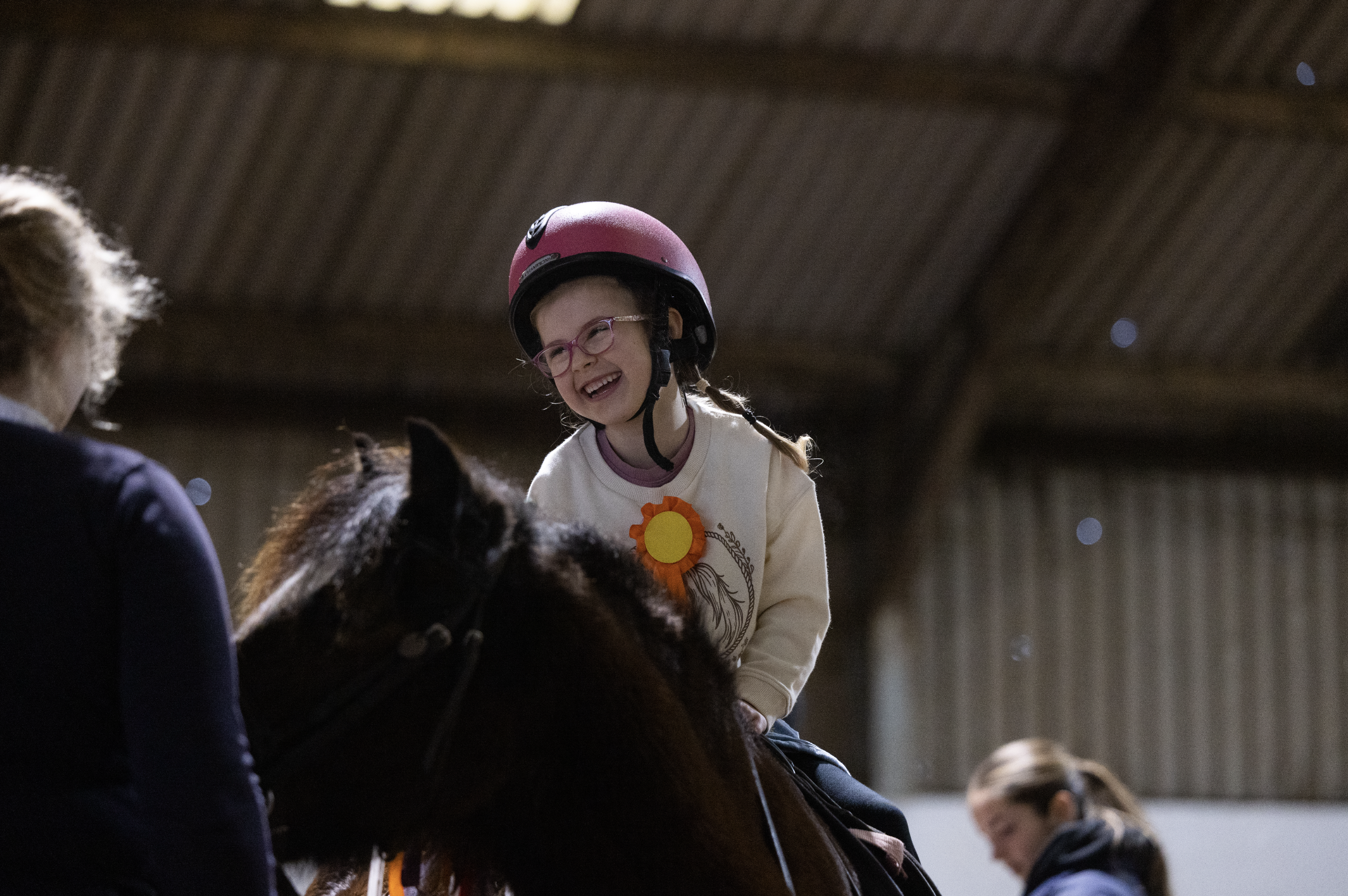 A young girl with glasses, wearing a pink helmet and a cream sweater with a yellow flower badge, is smiling and riding a horse indoors. An adult and another girl are nearby.