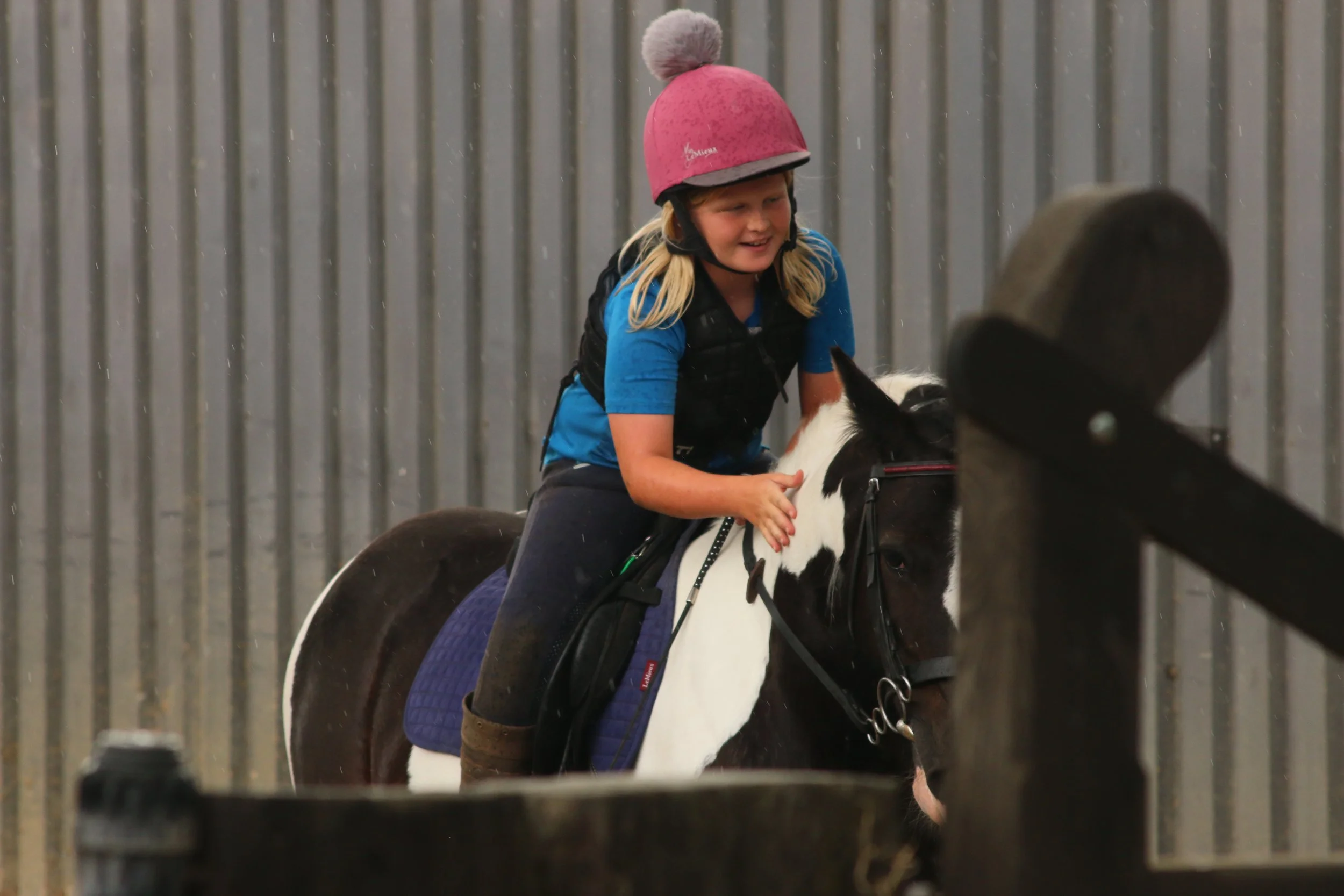 A young girl with blonde hair, wearing a pink helmet and blue shirt, laughing while riding a black and white horse in an indoor riding arena.