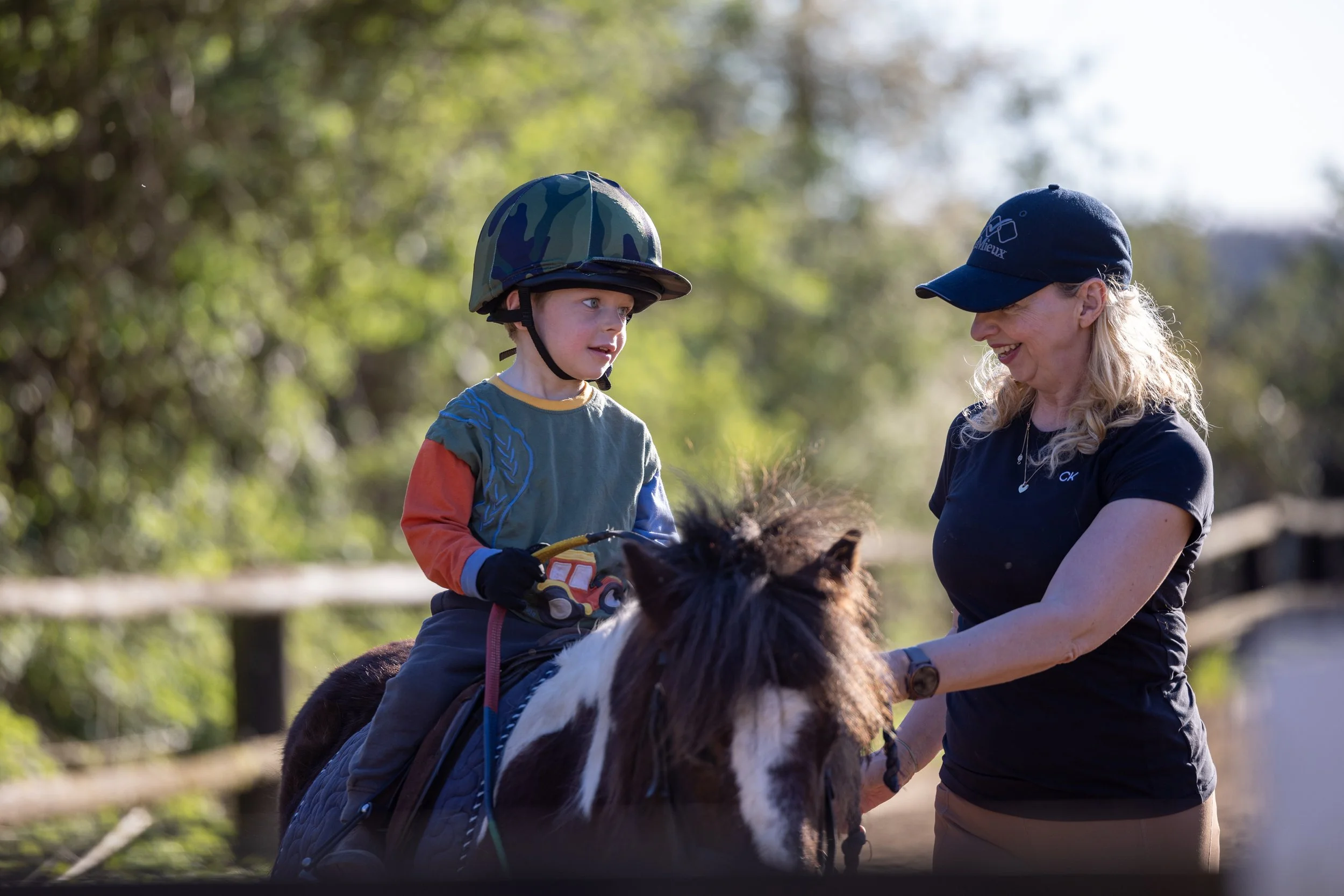 A young boy wearing a helmet and riding a pony, being assisted by a woman wearing a cap, in an outdoor setting with trees and a fence in the background.