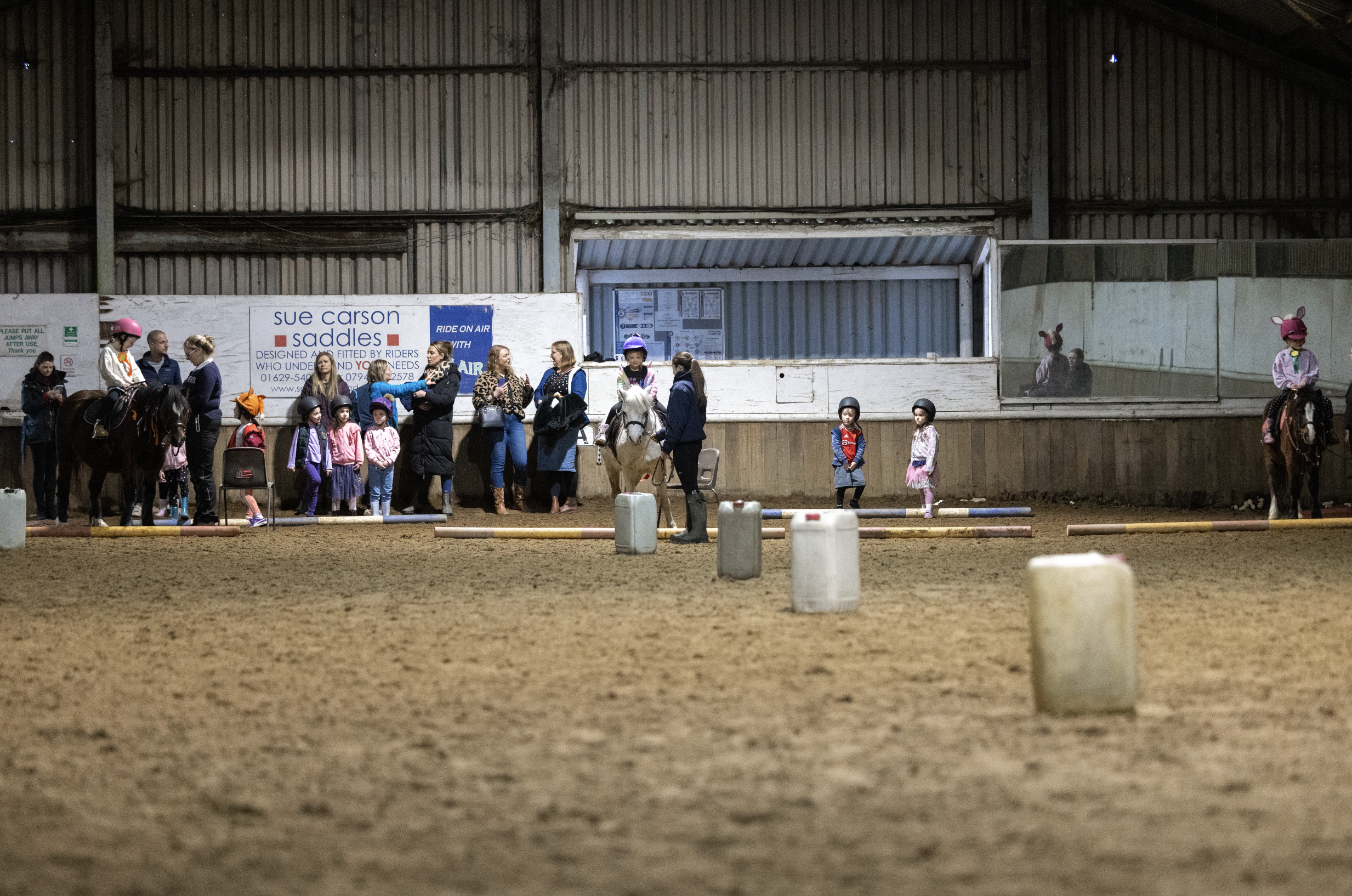 Children and adults in an indoor riding arena with horses, some children wearing helmets and pink bunny ears, lining up for a riding lesson or activity, with a banner that reads 'Sue Carson Saddles' in the background.