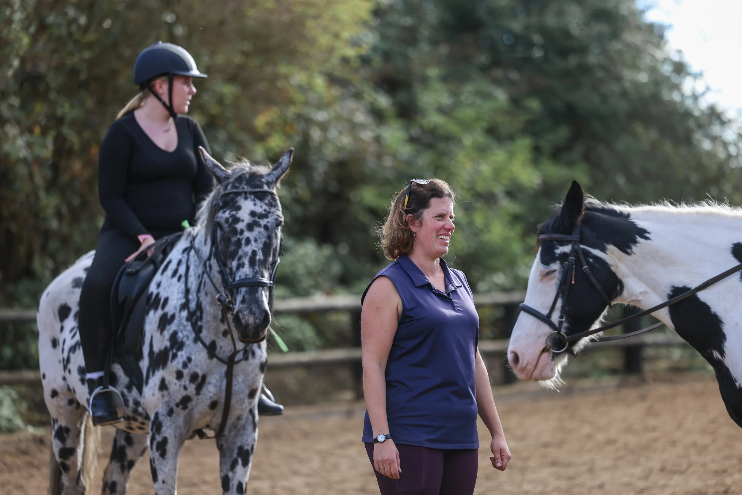 A woman riding a dappled gray horse stands next to another woman with a black and white spotted horse, both in an outdoor riding arena with trees in the background.