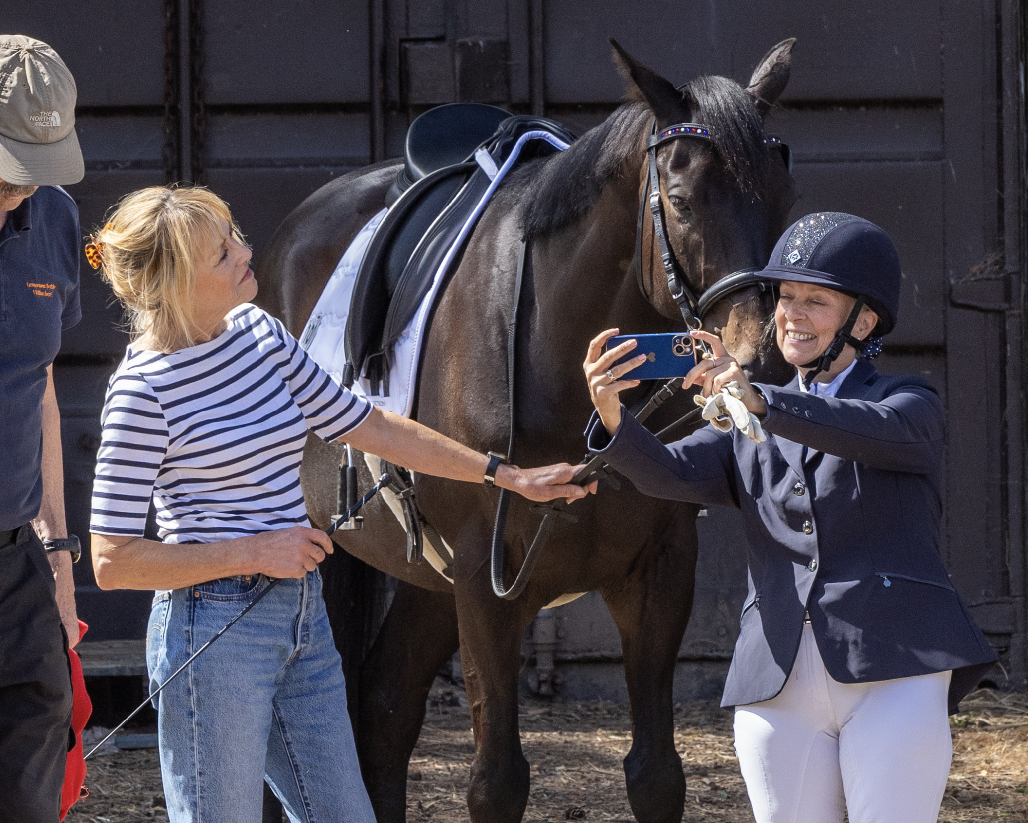A woman in a navy riding jacket and riding helmet taking a selfie with a smiling woman in a striped shirt beside a brown horse with a black mane, in a stable or horse yard.