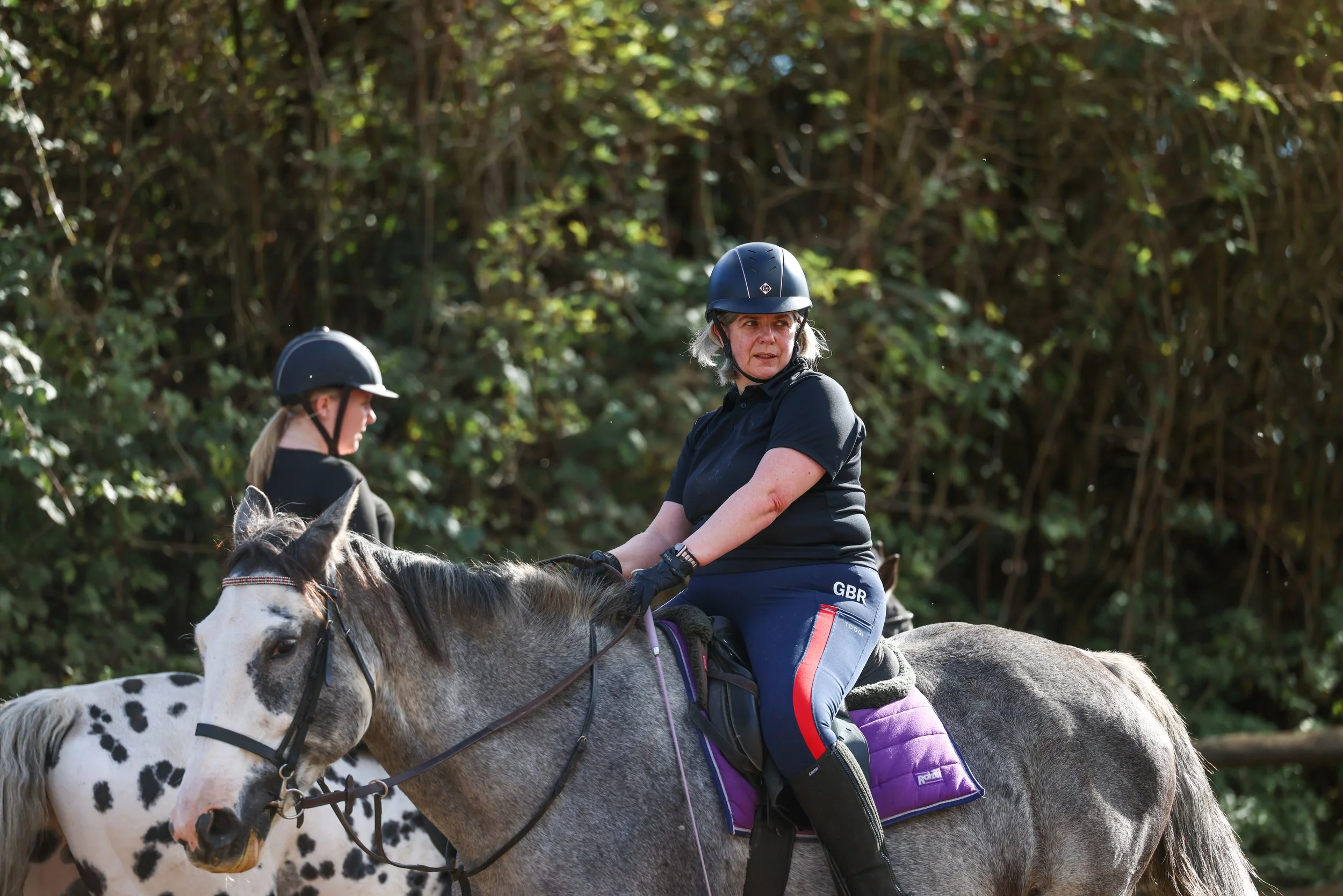 Two women riding horses outdoors, wearing black helmets and black shirts, with a background of dense green trees.