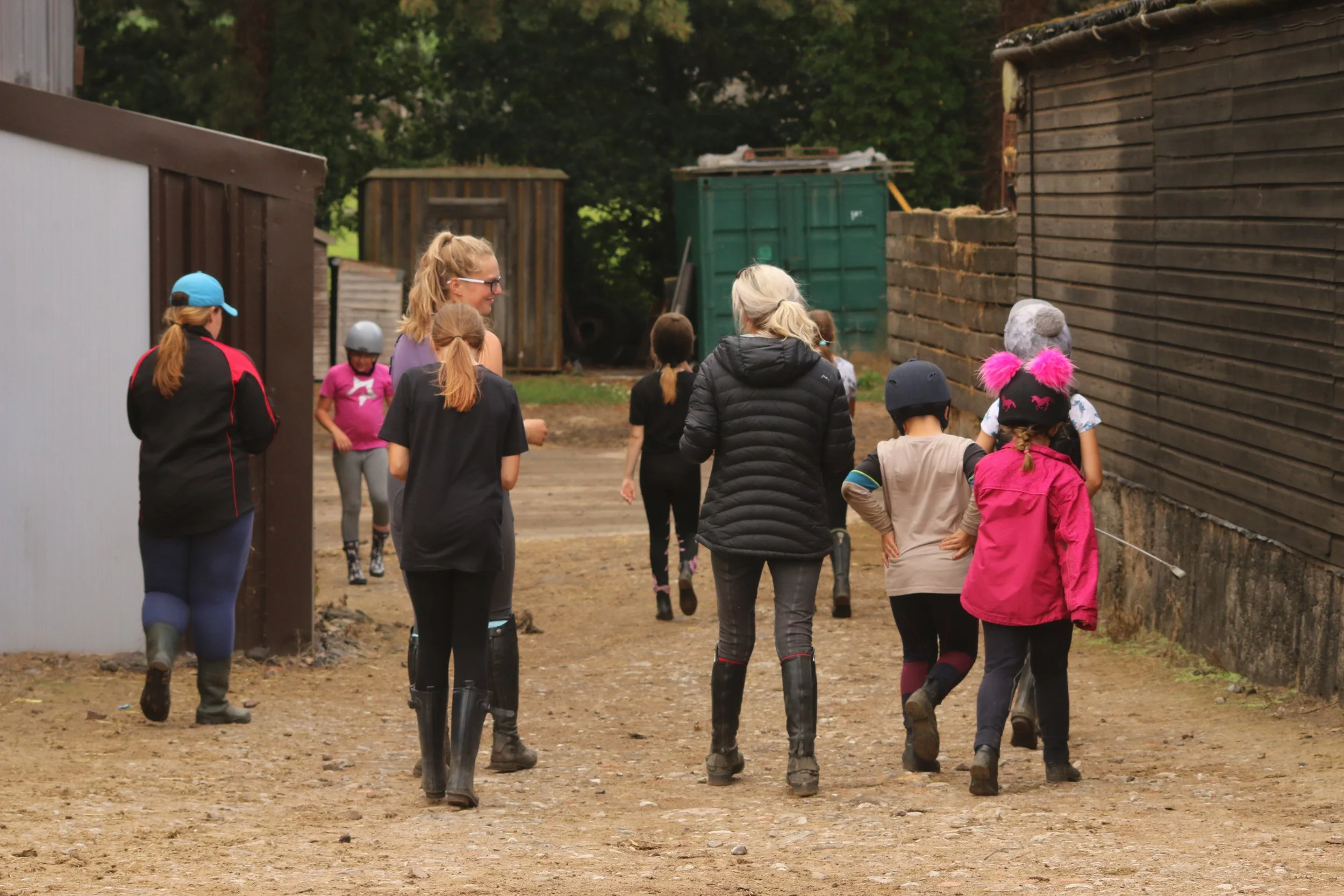 Children and adults in riding gear walking on a dirt path between wooden buildings at a farm or equestrian center.