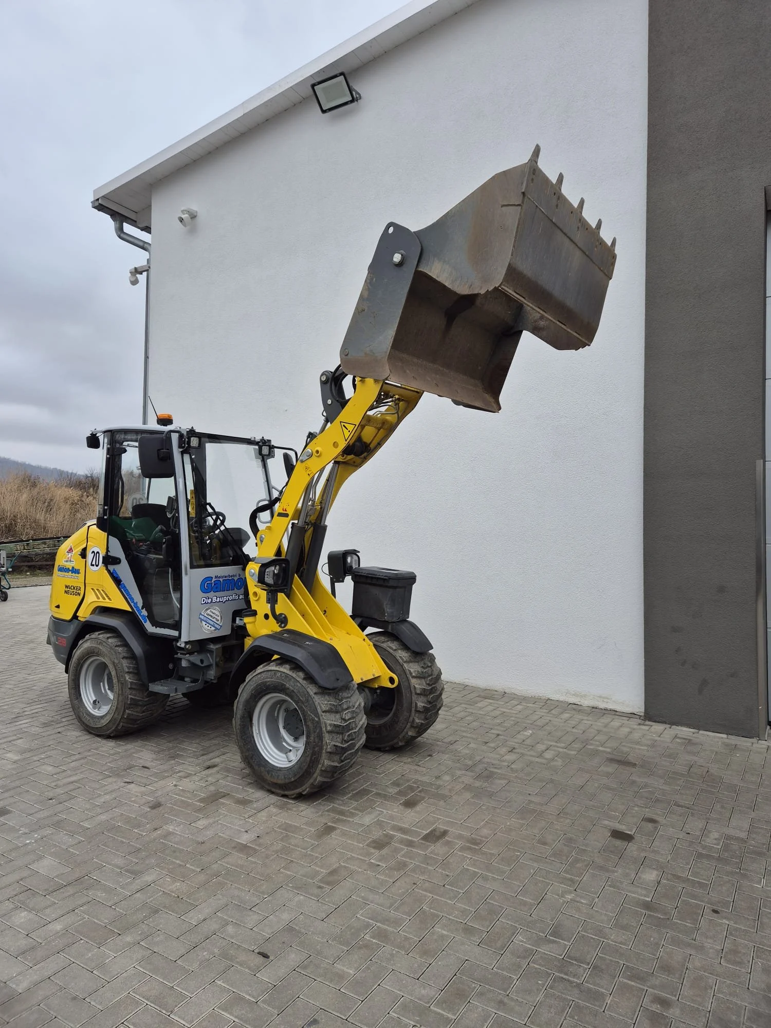 Kompakter gelber Baumaschinenlader mit großer Schaufel, auf einer gepflasterten Fläche vor einem weißen Gebäude mit grauer Wand zu sehen.