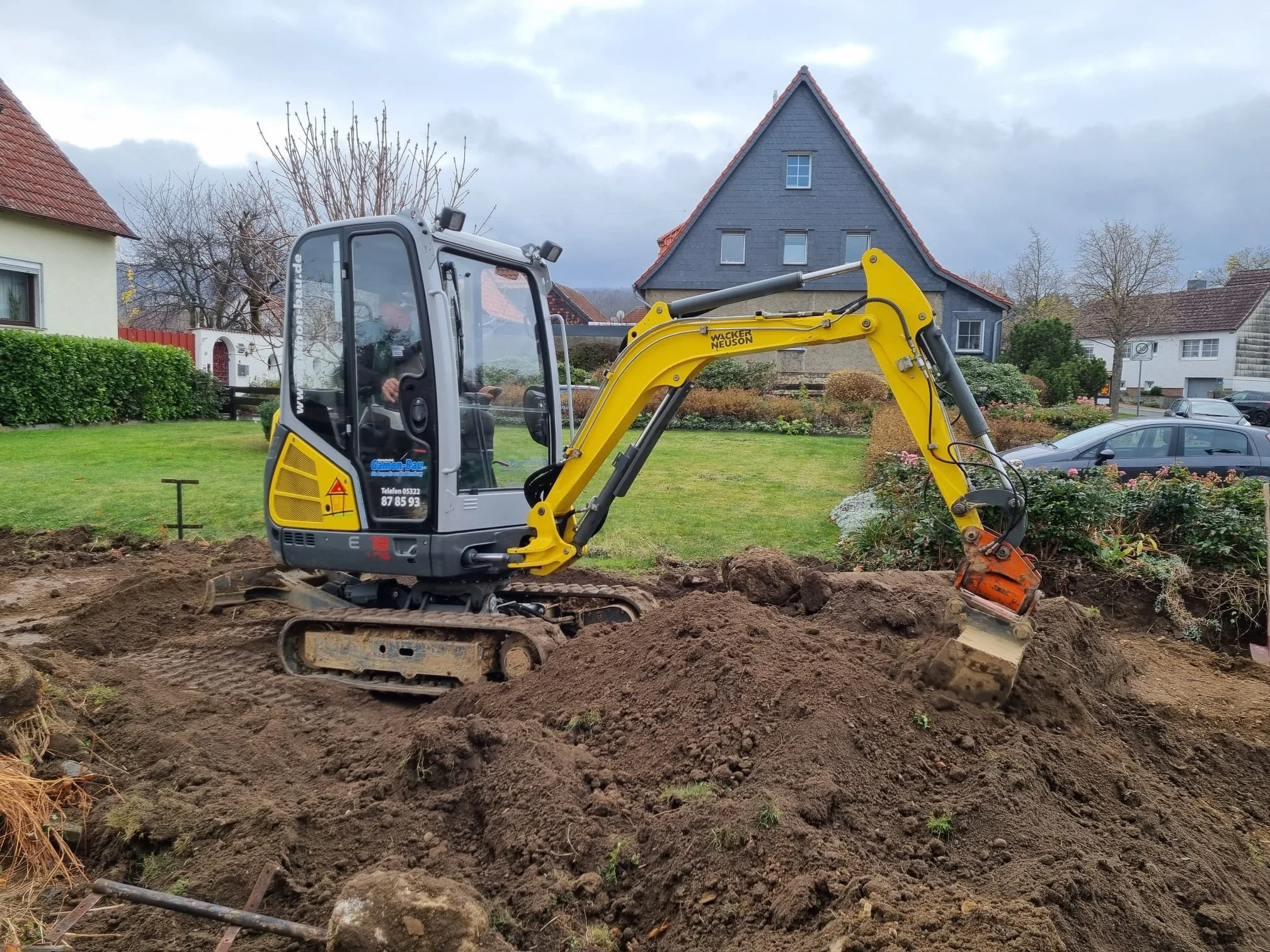 Kleiner Bagger, der auf einem Grundstück im Erdreich arbeitet, um Erde zu bewegen, mit einem Haus im Hintergrund und einem bewölkten Himmel.