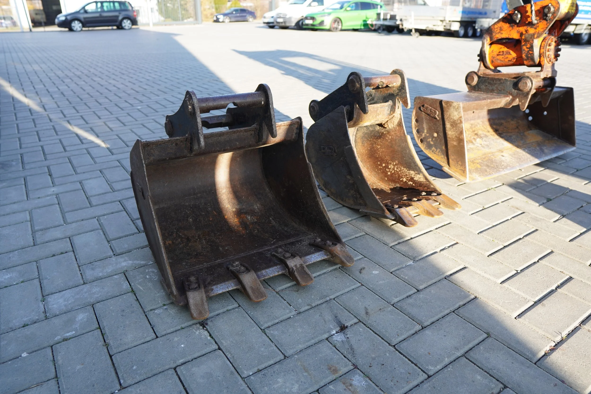 Drei Bagger-Schaufeln auf einer gepflasterten Fläche im Freien, mit Autos im Hintergrund.