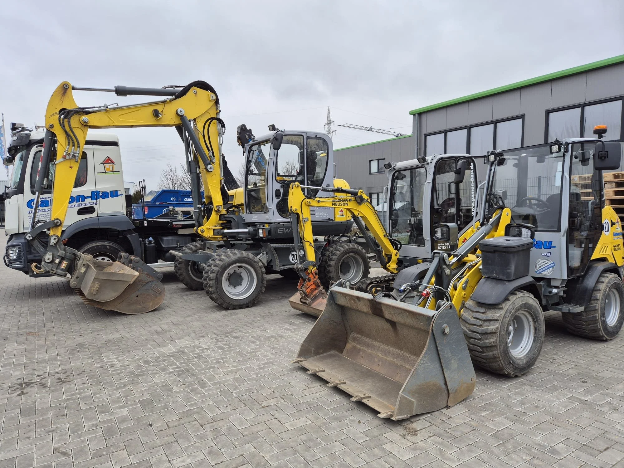 Verschiedene Baumaschinen, darunter Minibagger und Radlader, vor einer Fabrik im Freien. Von GMS | Gamon Miet-Service