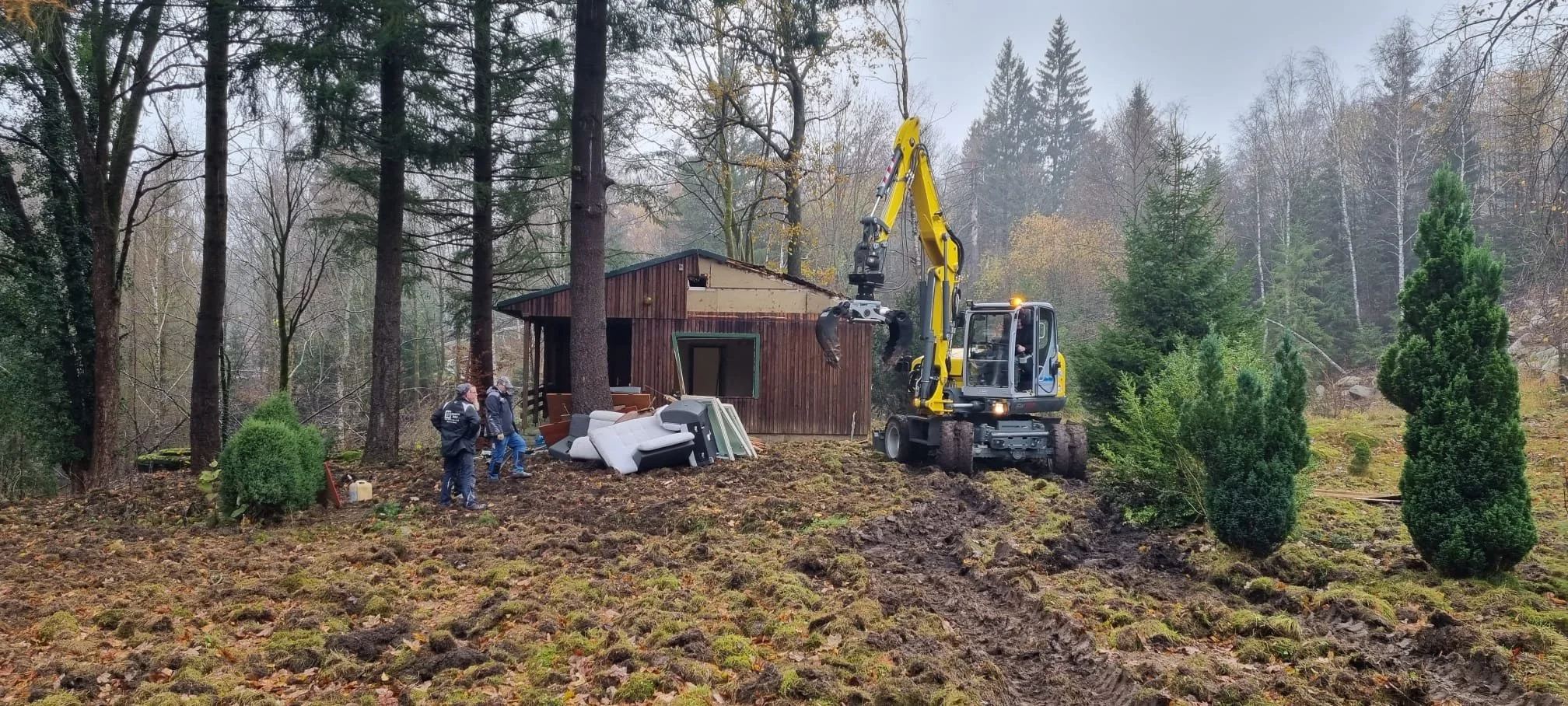 Bergwerk bei der Arbeit: Bauarbeiter im Wald neben einem kleinen Holzhaus, Müll auf dem Boden, ausgefahrene Erde und Einsatz eines Mobilbaggers