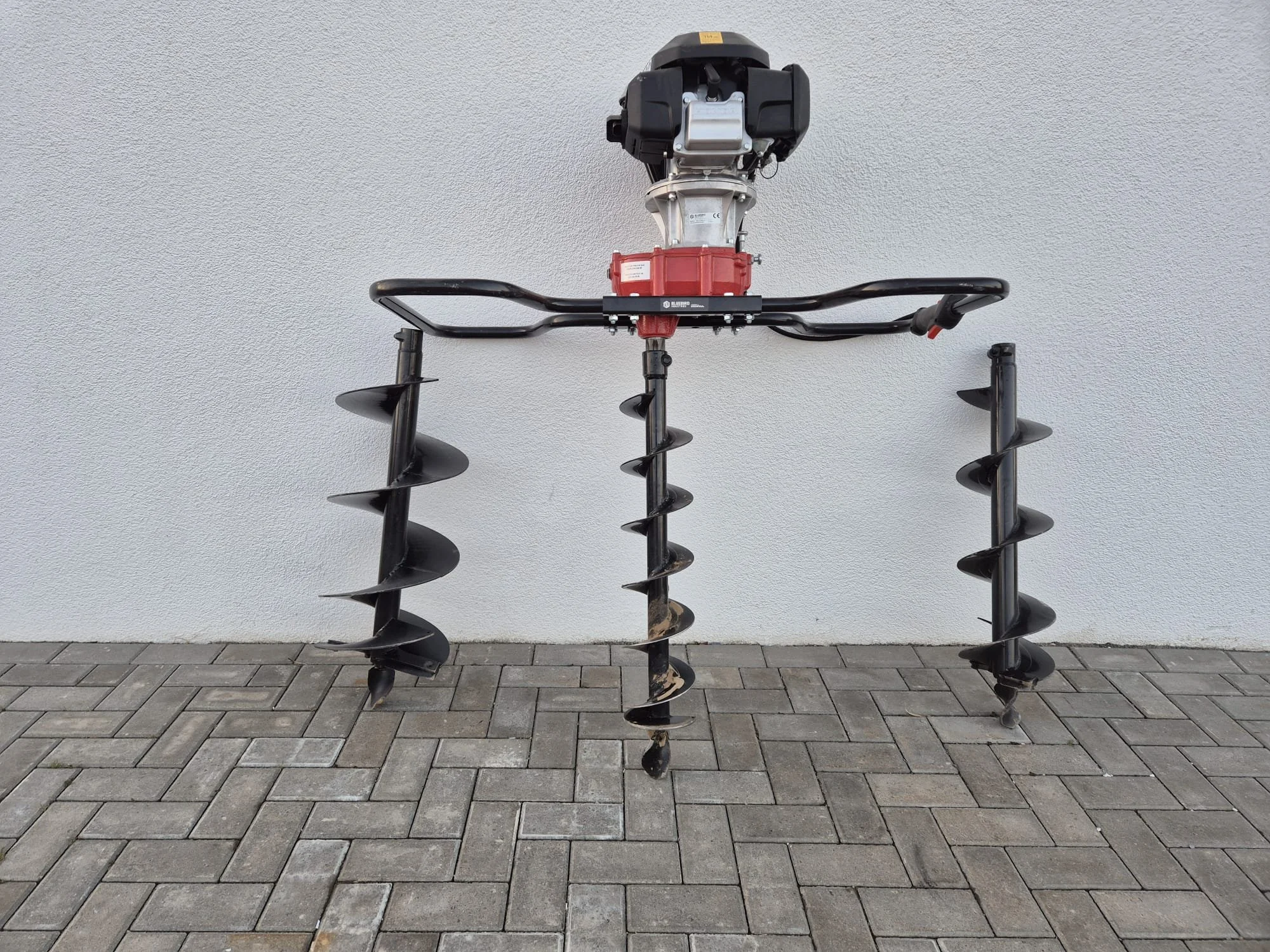 Motorsäule mit drei Bohrern in verschiedenen Größen, auf einer Pflastersteinfläche vor einer grauen Wand.