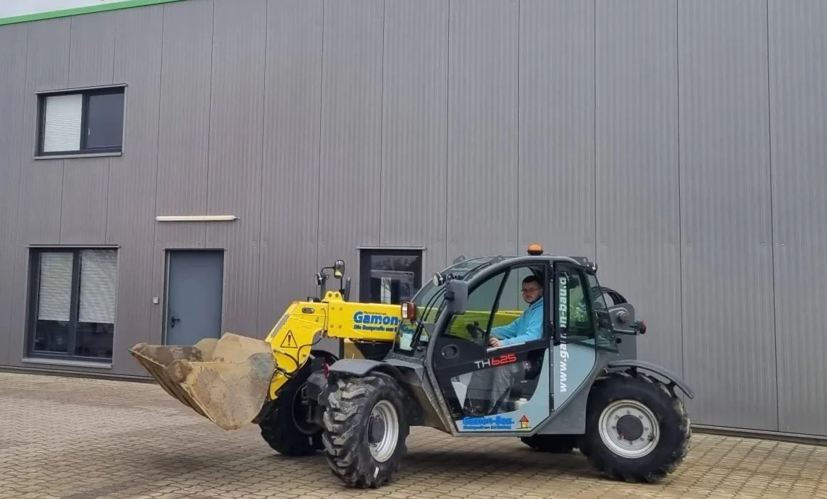 Ein Mann sitzt in einem Teleskoplader mit Ballenzange, vor einer modernen, grauen Gebäude. Das Fahrzeug trägt die Beschriftung 'Gamon' und hat einen gelben Anbau.