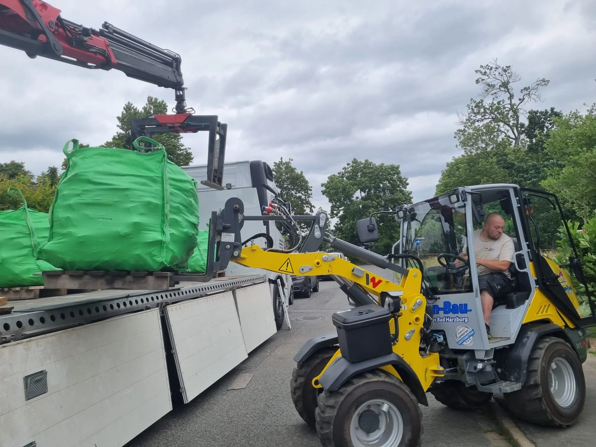 Mann bedient einen Radlader, der große grüne Säcke auf einen Lastwagen hebt, auf einer Straße, umgeben von Bäumen, bei bewölktem Himmel.