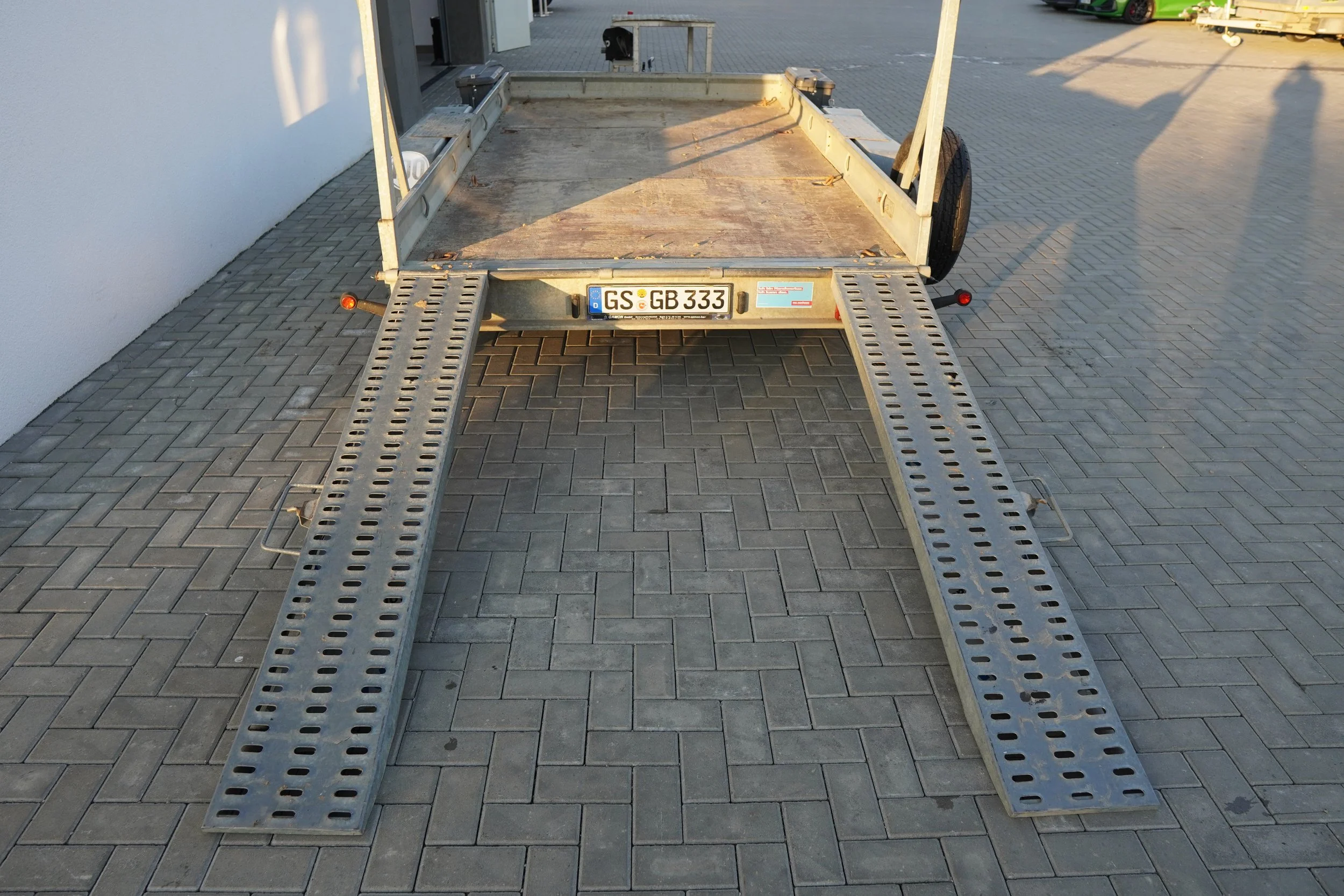 Ein leerer Maschinentransporter mit Rampe auf gepflastertem Boden, im Hintergrund Gebäude und Schatten.