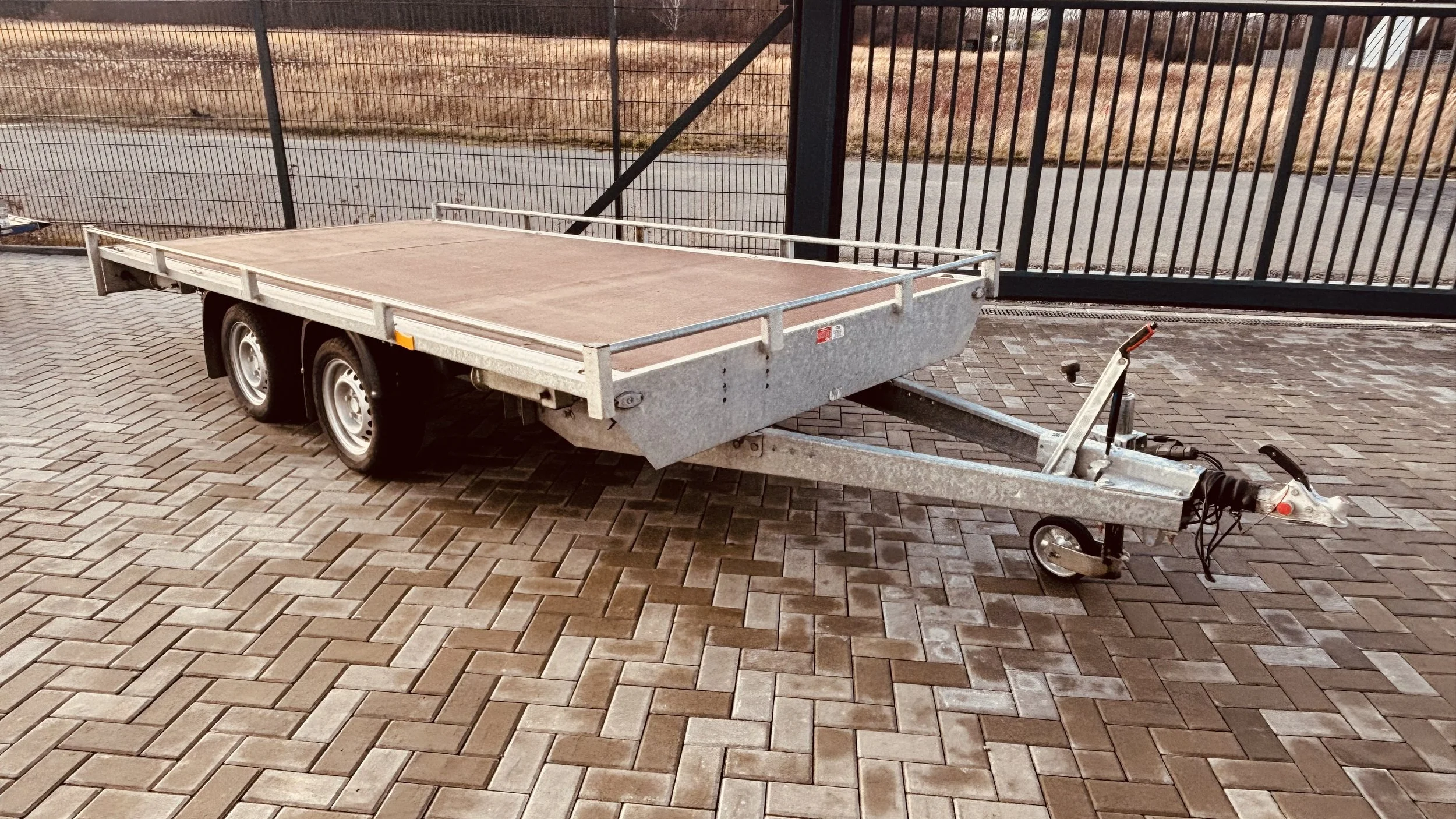 Leerer Anhänger mit Holzladefläche und einer Zugvorrichtung auf einem gepflasterten Boden, im Hintergrund eine Metalltor und eine landwirtschaftlich anmutende Umgebung.