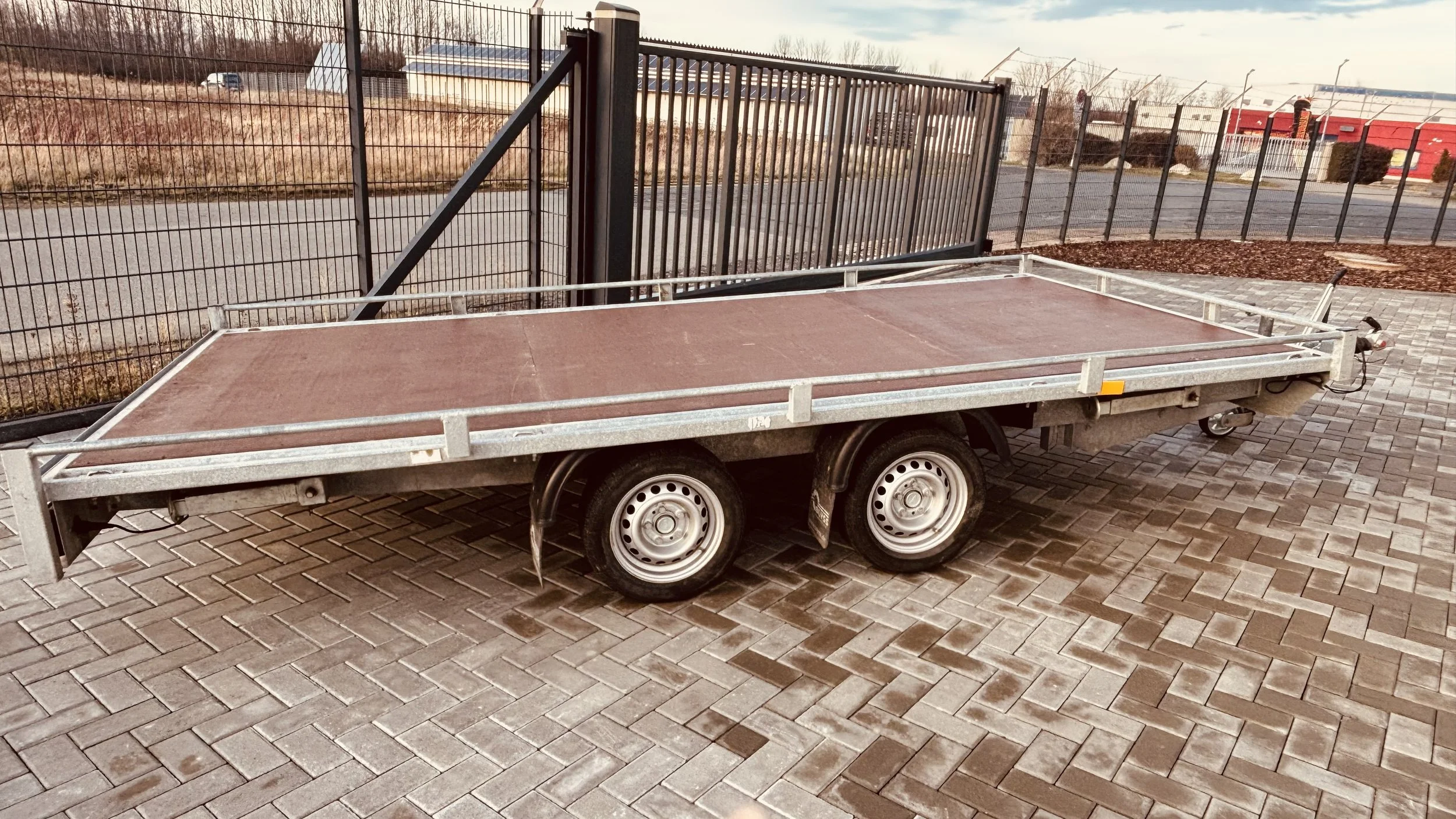 Ein leerer, flacher Autotrailer auf einem gepflasterten Boden neben einem Zaun, vermutlich für den Transport von Gütern oder Fahrzeugen.