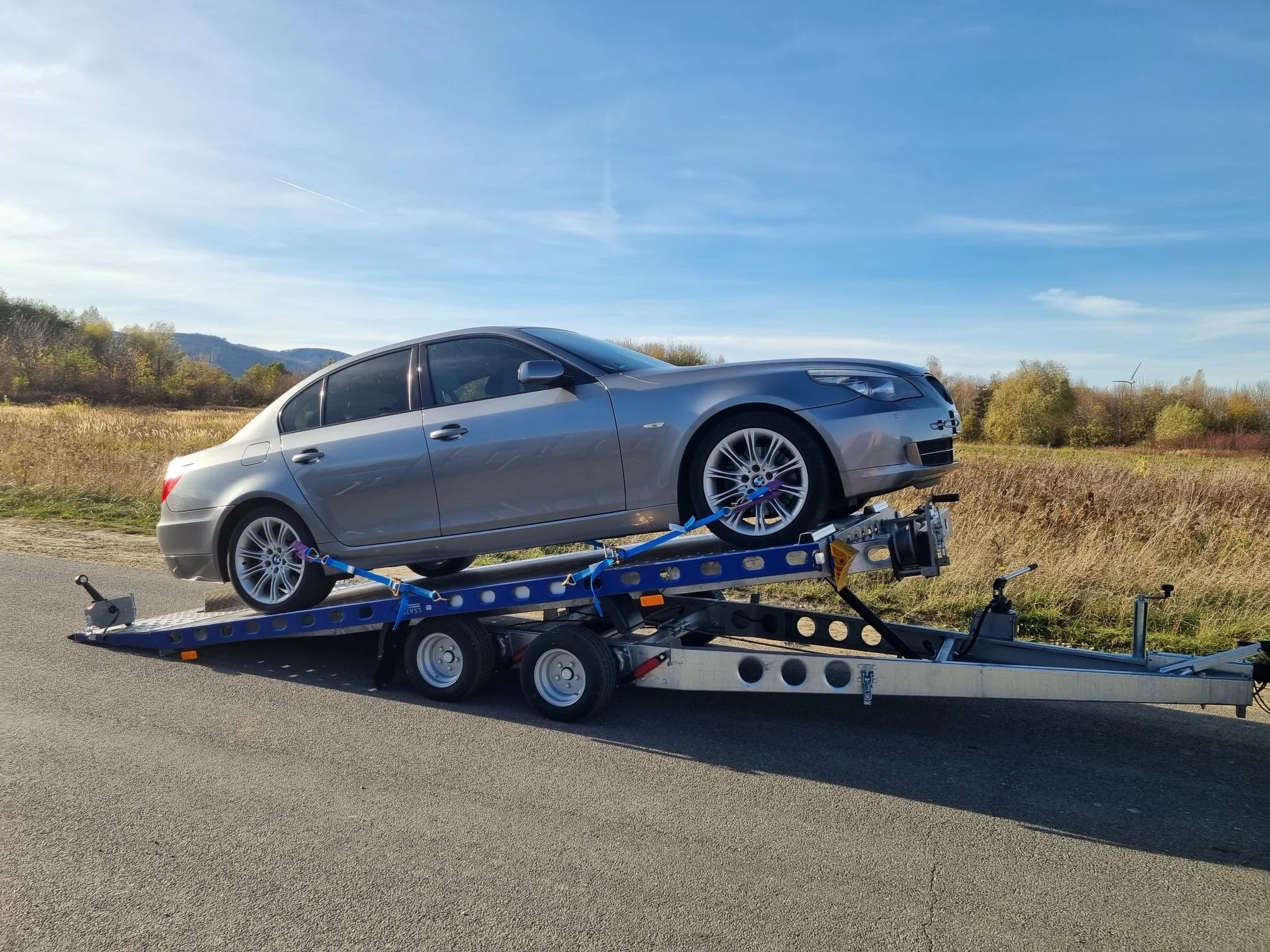 Ein silberner Pkw auf einem Autotrailer auf einer Landstraße bei sonnigem Himmel.