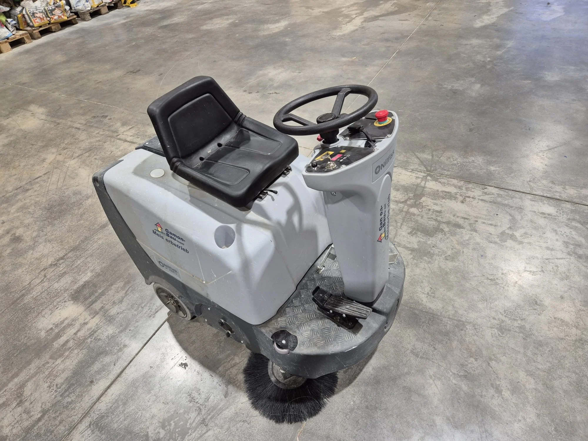 Eine graue Kehrmaschine mit Sitz und Lenkrad auf einer glatten, grauen Bodenfläche in einem Lagerraum