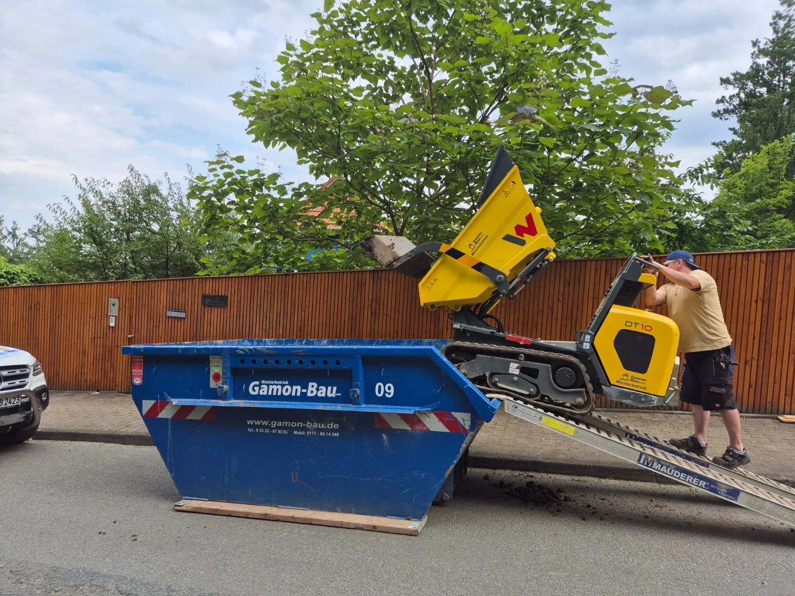 Ein Mann bedient einen Raupendumper, um Erde in einen großen blaue Container zu kippen. Es ist Tag und im Hintergrund sind Bäume und ein Holzzaun zu sehen.