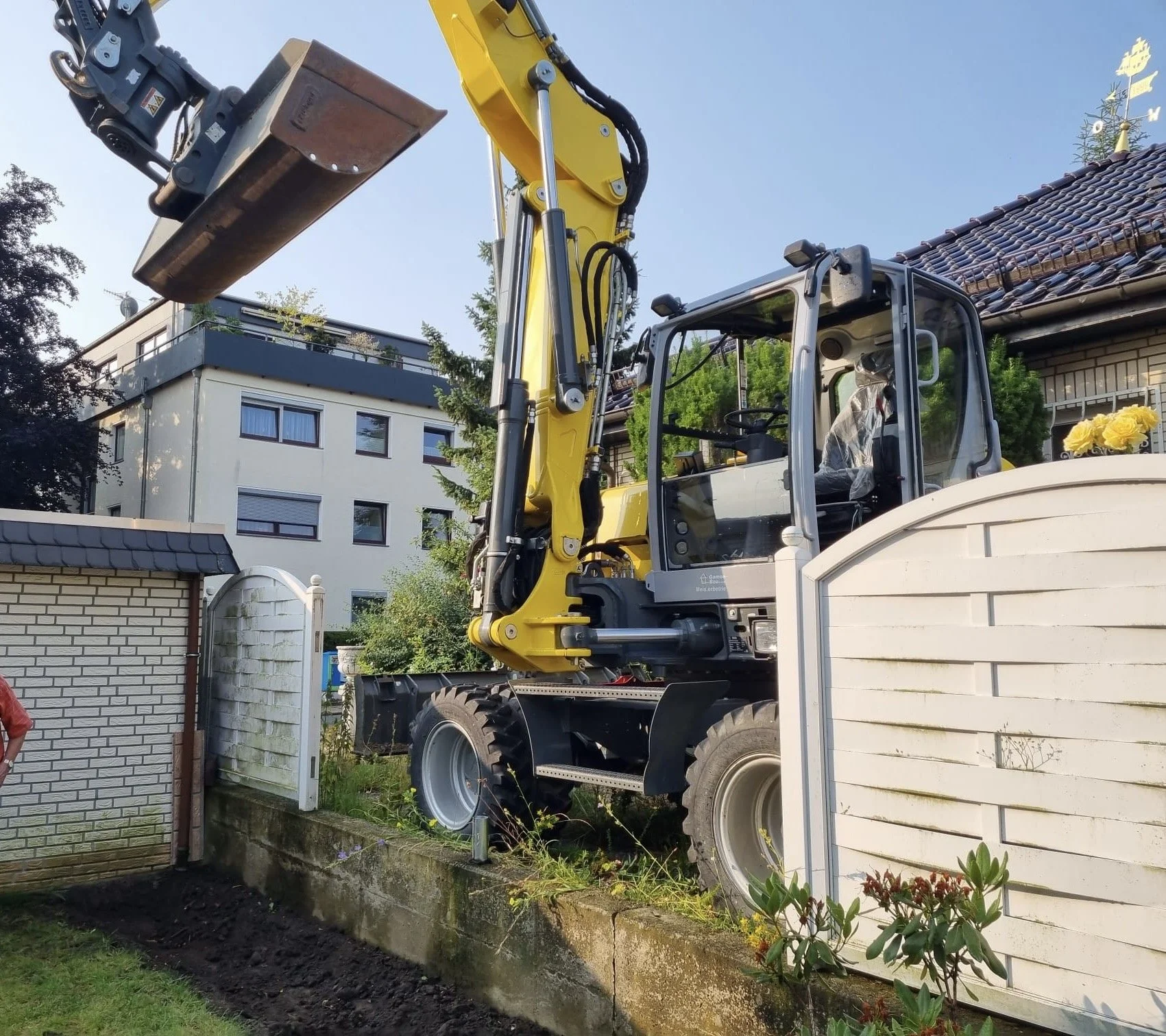 Ein gelber Mobilbagger hebt einen Baggerarm mit Anbau über eine weiße Einfriedung in einem Garten. Im Hintergrund sind Wohngebäude und Bäume zu sehen.