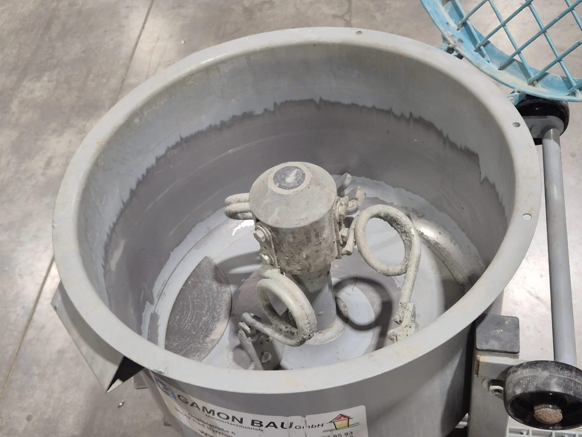 Leerer Mörtelmischbehälter mit verstaubtem, zerlegtem Rührgerät in der Mitte, auf dem Boden liegt ein Metallspachtel, im Hintergrund eine blaue Wanne, alles steht auf einem Fliesenboden.