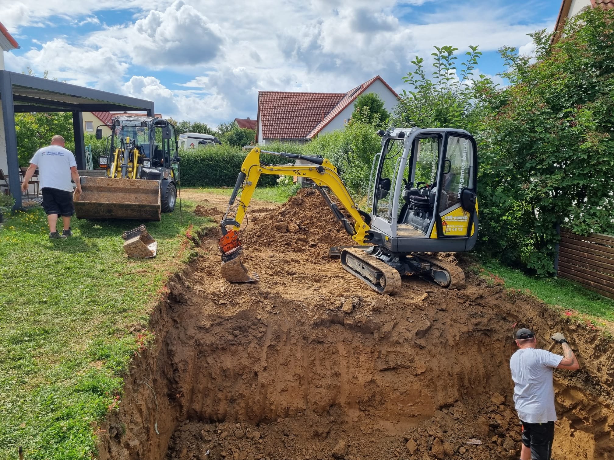 Bauarbeiter arbeiten an einer Baugrube mit einem kleinen Bagger, während andere Arbeiter auf einer Wiese sind. Es ist ein sonniger Tag mit einigen Wolken am Himmel.