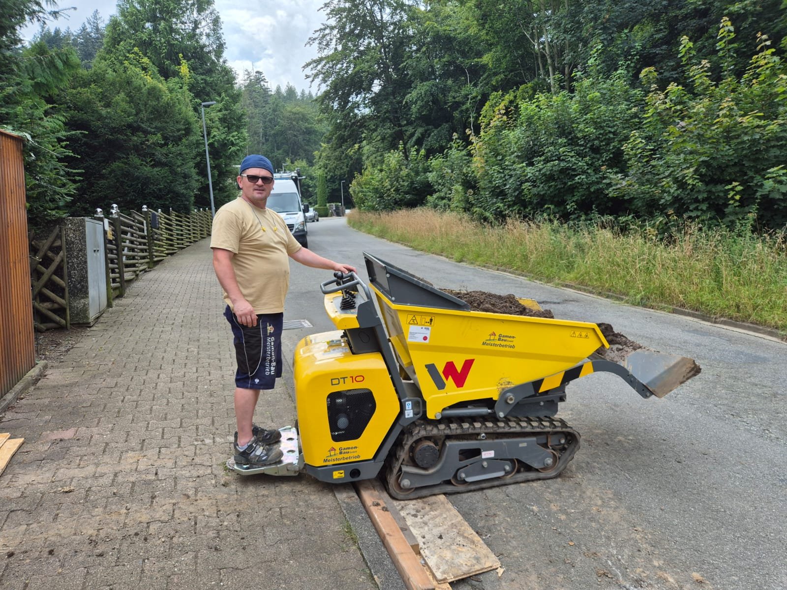 Ein Mann steht auf dem Raupendumper auf einer Straße, die gleichzeitig bergauf und ab führt, mit Bäumen und einem weißen Van im Hintergrund.