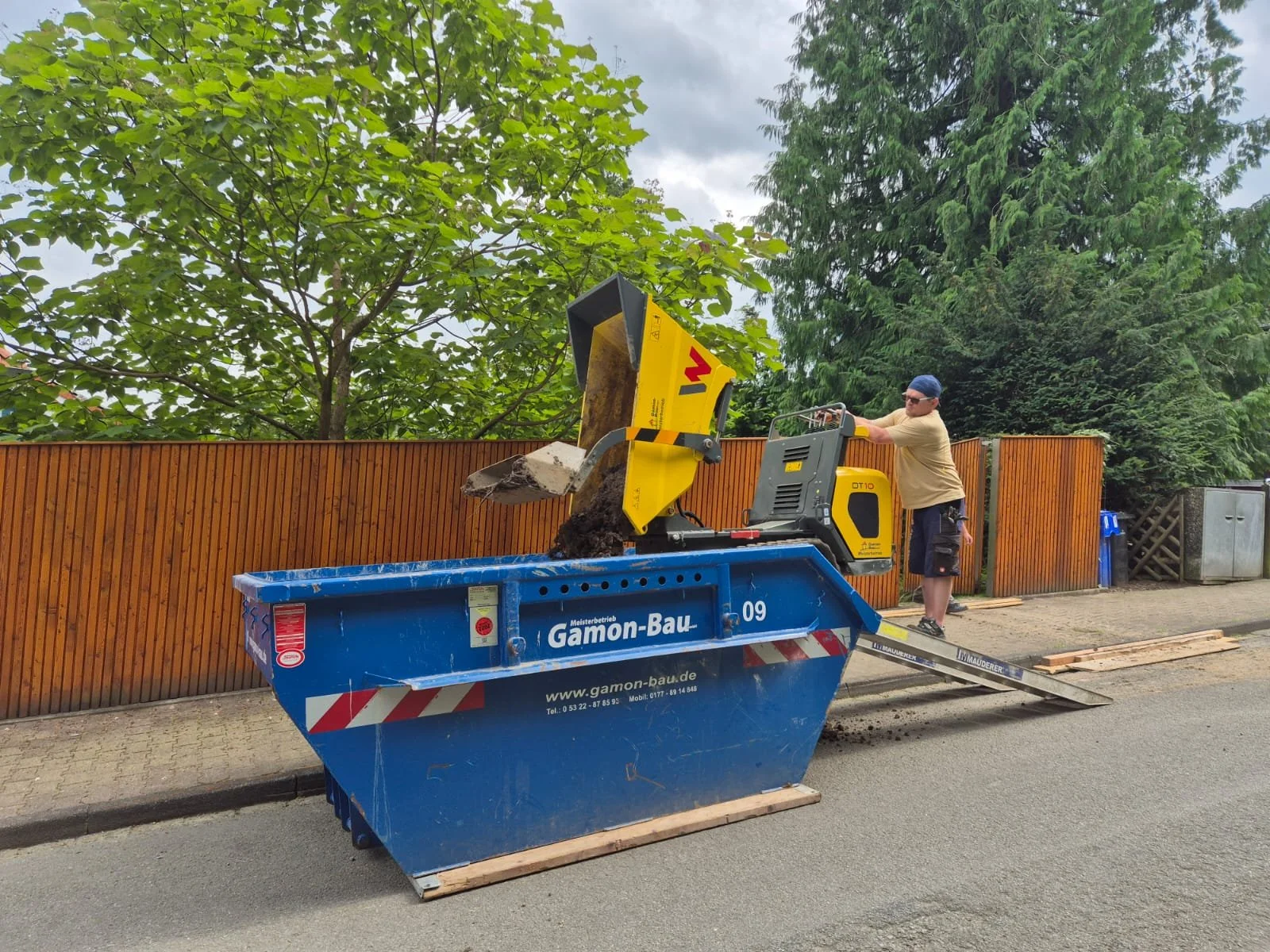 Ein Mann bedient einen gelben Raupendumper, die Erde in einen blauen Container auf der Straße wirft, mit einem Holzsteg, der den Container mit dem Straßenrand verbindet, im Hintergrund ein Holzzaun, Bäume und ein grauer Himmel.