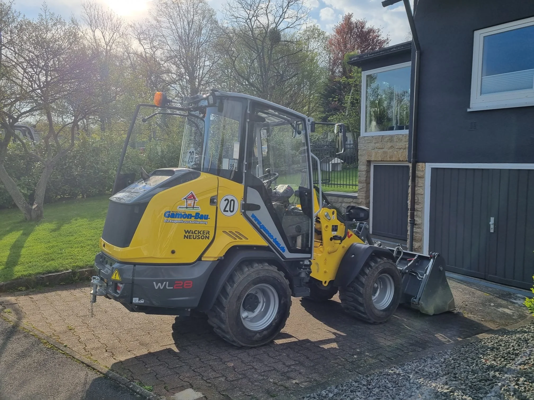 Radlader mit geschlossener Kabine, parkt auf einer gepflasterten Fläche vor einem schwarzen Haus mit weißem Fenster, im Hintergrund grüne Bäume und ein Himmel mit Wolken.