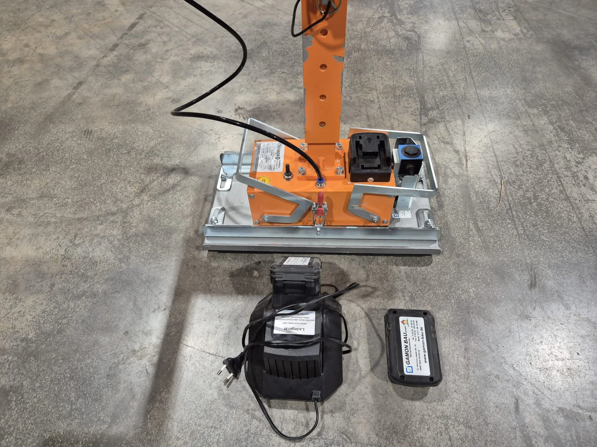 Gerät zur Bodenverankerung, bestehend aus einer Metallbasis, orangefarbenem Pfosten, elektrischen Komponenten und einem externen Netzteil auf einem Betonboden.