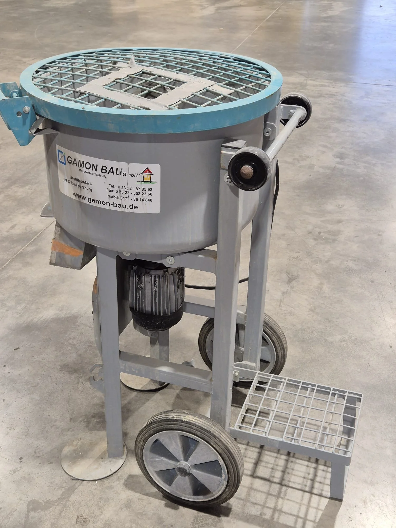 Ein grauer Bauwagen mit einem blauen Rand und einem Gitterdeckel, stehen auf einem Betonboden. Das Bauwagen trägt ein Aufkleber mit Kontaktinformationen und einer Webseite.