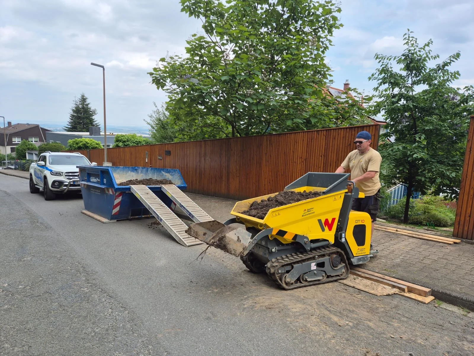 Mit einem kleinen Baumaschinen, die Erde bewegt, arbeitet ein Mann auf einer Straße neben einem gelben Transportgerät. Im Hintergrund sind Bäume, eine Holzzäune und ein parkendes Auto zu sehen.