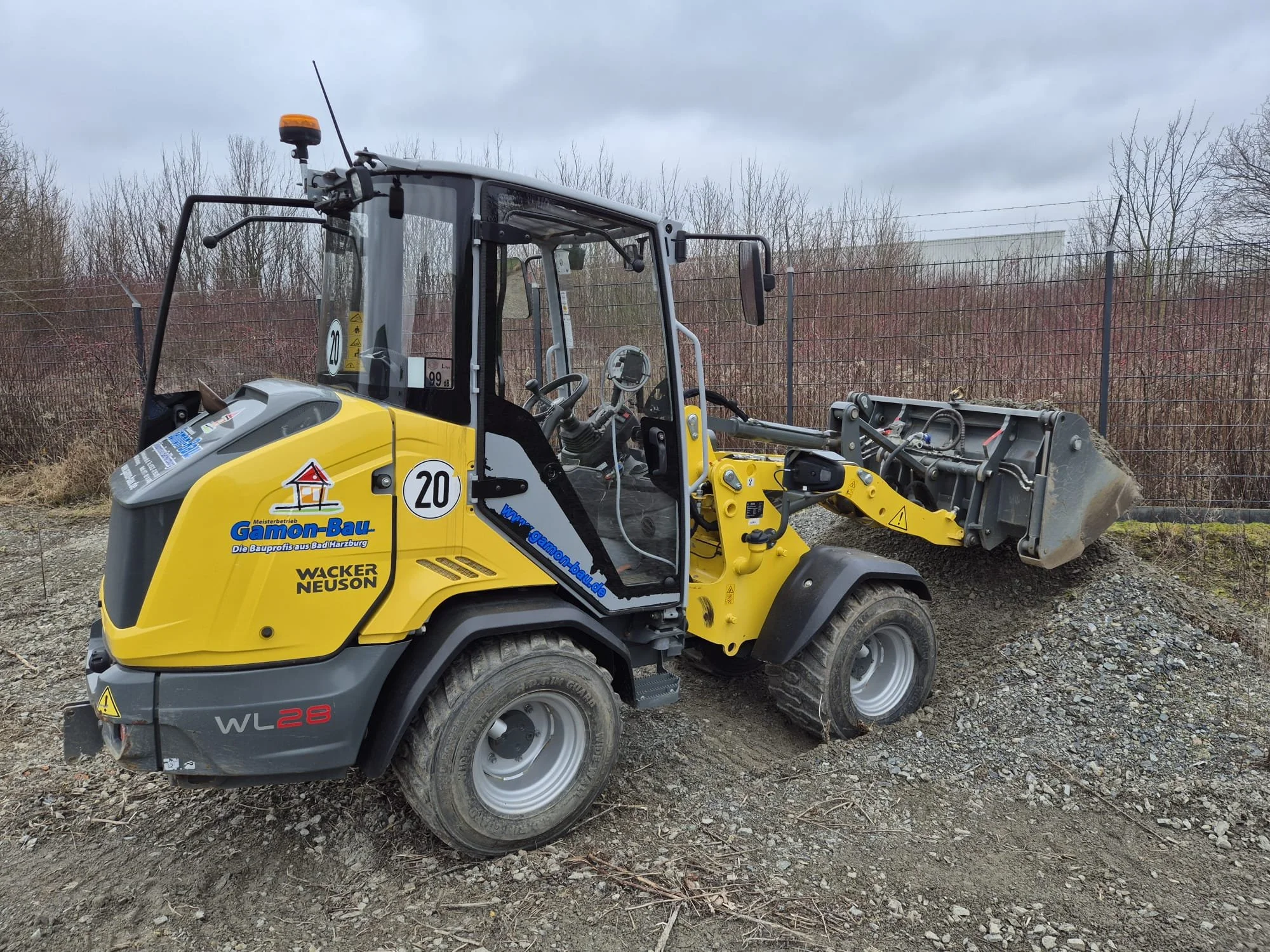 Gelbes Baustellenfahrzeug (Radlader) mit Schaufel an der Front, auf einer Baufläche mit Steinen und Erde, im Hintergrund Drahtzaun und Bäume bei bewölktem Himmel.