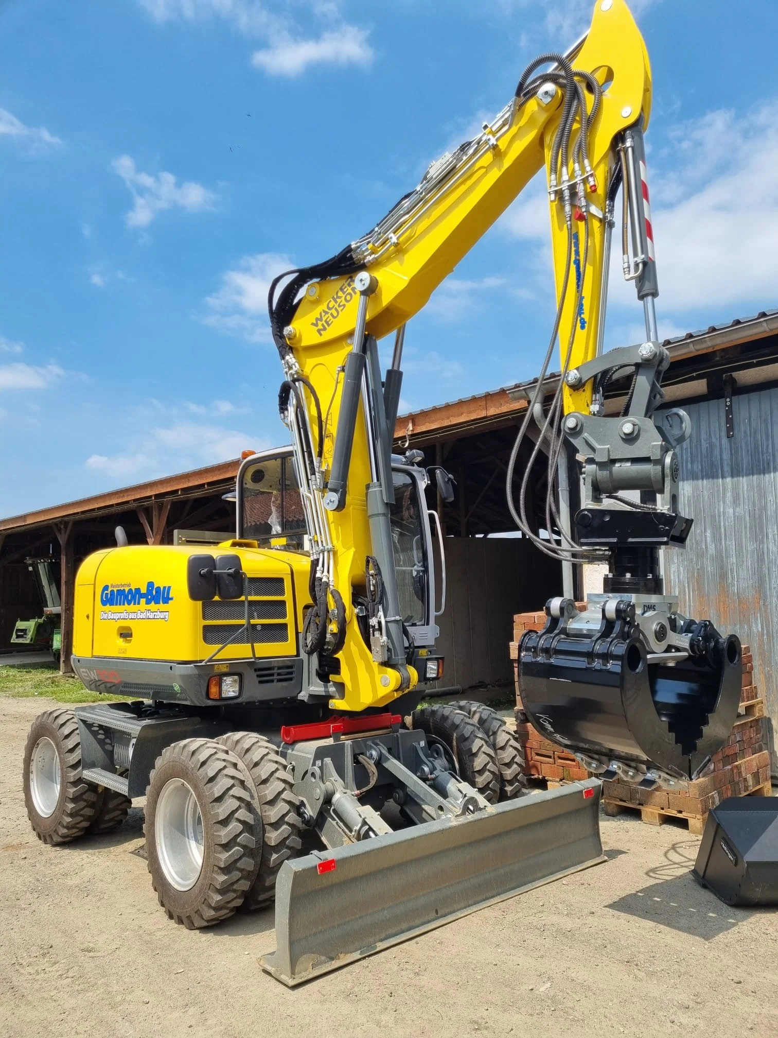 Gelber Mobilbagger von Gamon mit Hydraulik Arm und Frontblatt, auf Baustelle mit Holzstapeln im Hintergrund.