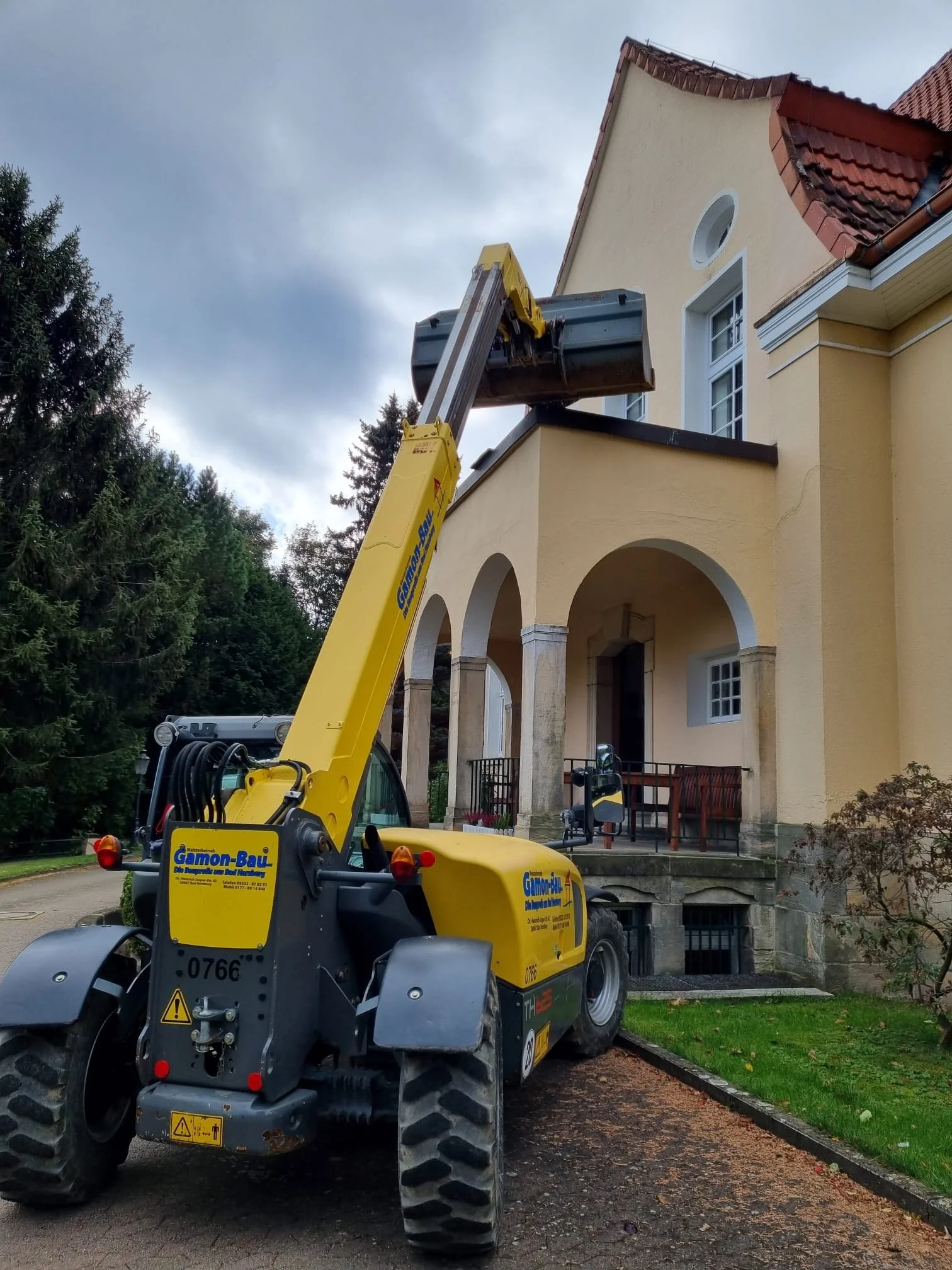 Baufahrzeug mit Teleskoplader hebt eine Dachschindel an einem mehrstöckigen, gelben Haus mit rotem Dach, um Reparaturen durchzuführen.