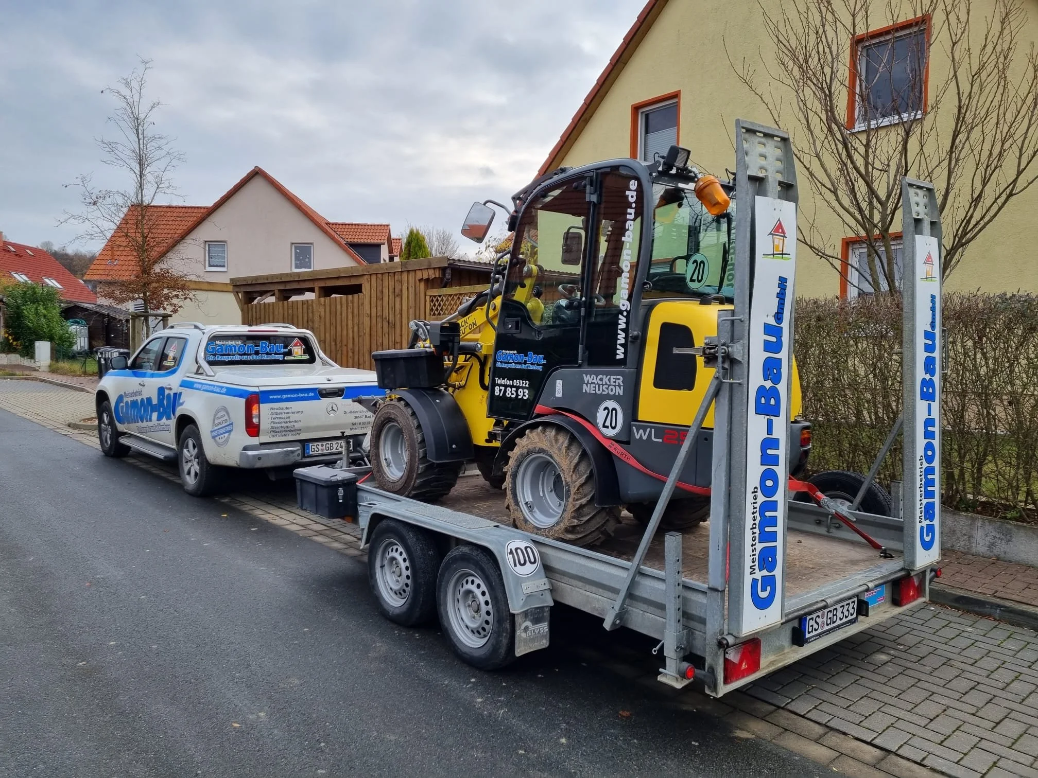 Ein Baufahrzeug mit einem kleinen Bagger auf einem Anhänger parkt auf der Straße vor einem Haus, während ein weißes Firmenfahrzeug daneben steht.