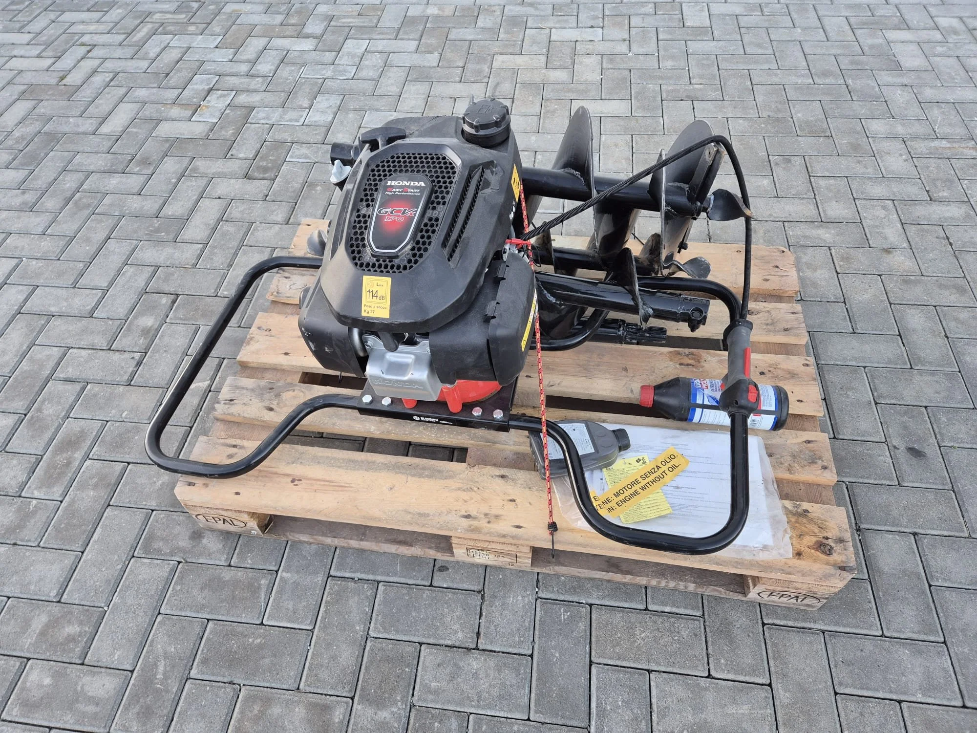 Ein motorisiertes Handbohrgerät auf einer Holzpalette, mit Benzinmotor, Griffen, und Anweisungen in mehreren Sprachen.