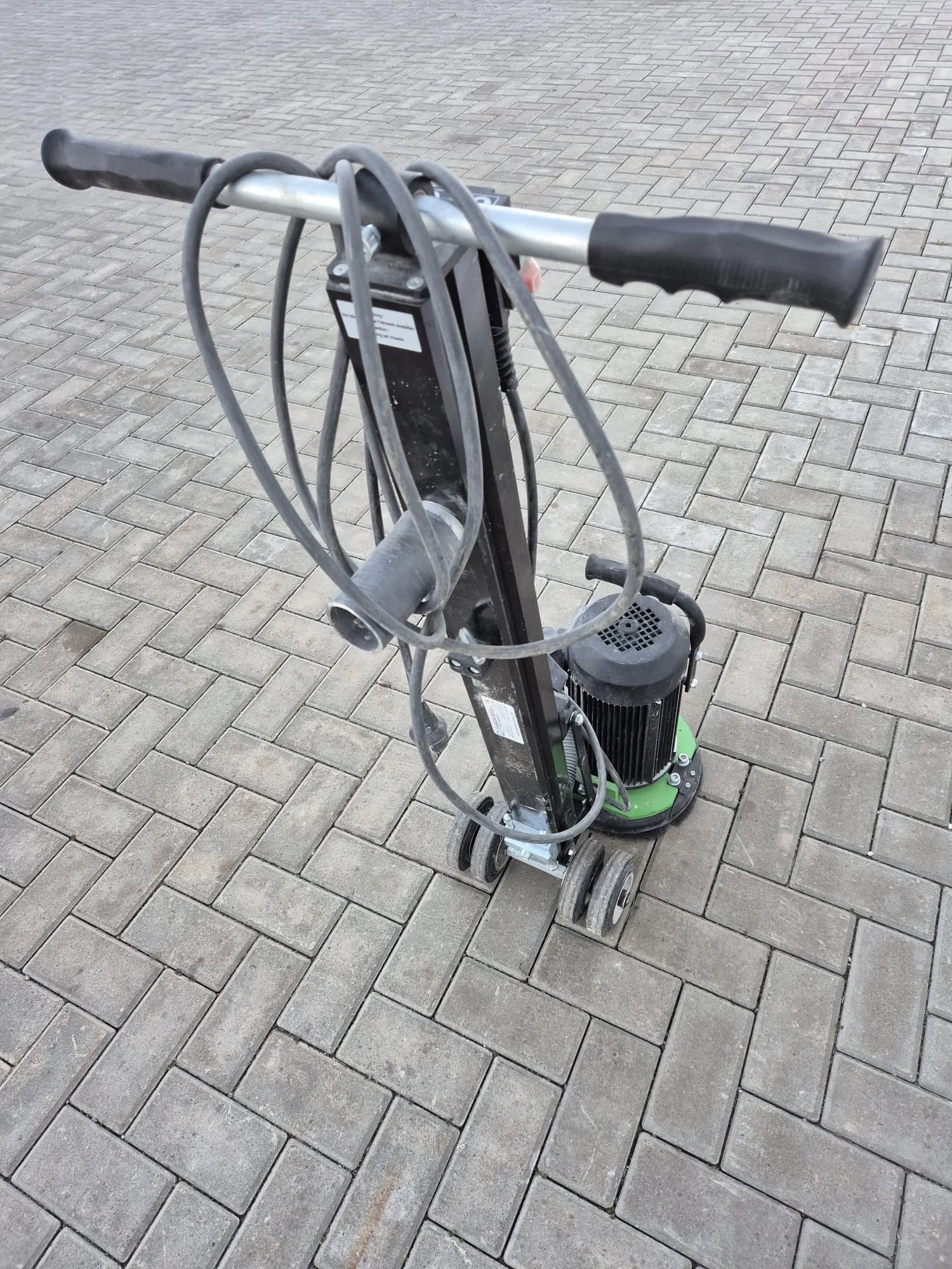Ein elektrisches Bodenreinigungsgerät auf gepflastertem Boden, mit Griffen und Kabel.