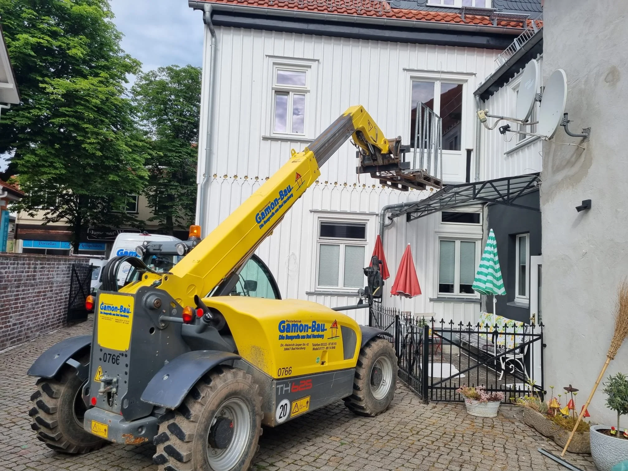 Ein gelber Teleskoplader heben auf eine Terrasse vor einem weißen Haus. Das Haus hat weiße Fenster und Satellitenschüsseln, sowie einen überdachten Balkon. Im Garten sind bunte Sonnenschirme und Pflanzen.