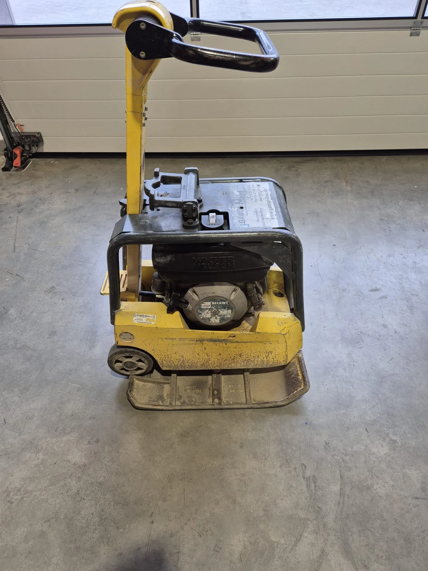 Gelbe Vibrationsplatte mit einem schwarzen Motor, Griff und Knöpfen, auf einer Betonboden in einer Garage oder Werkstatt.