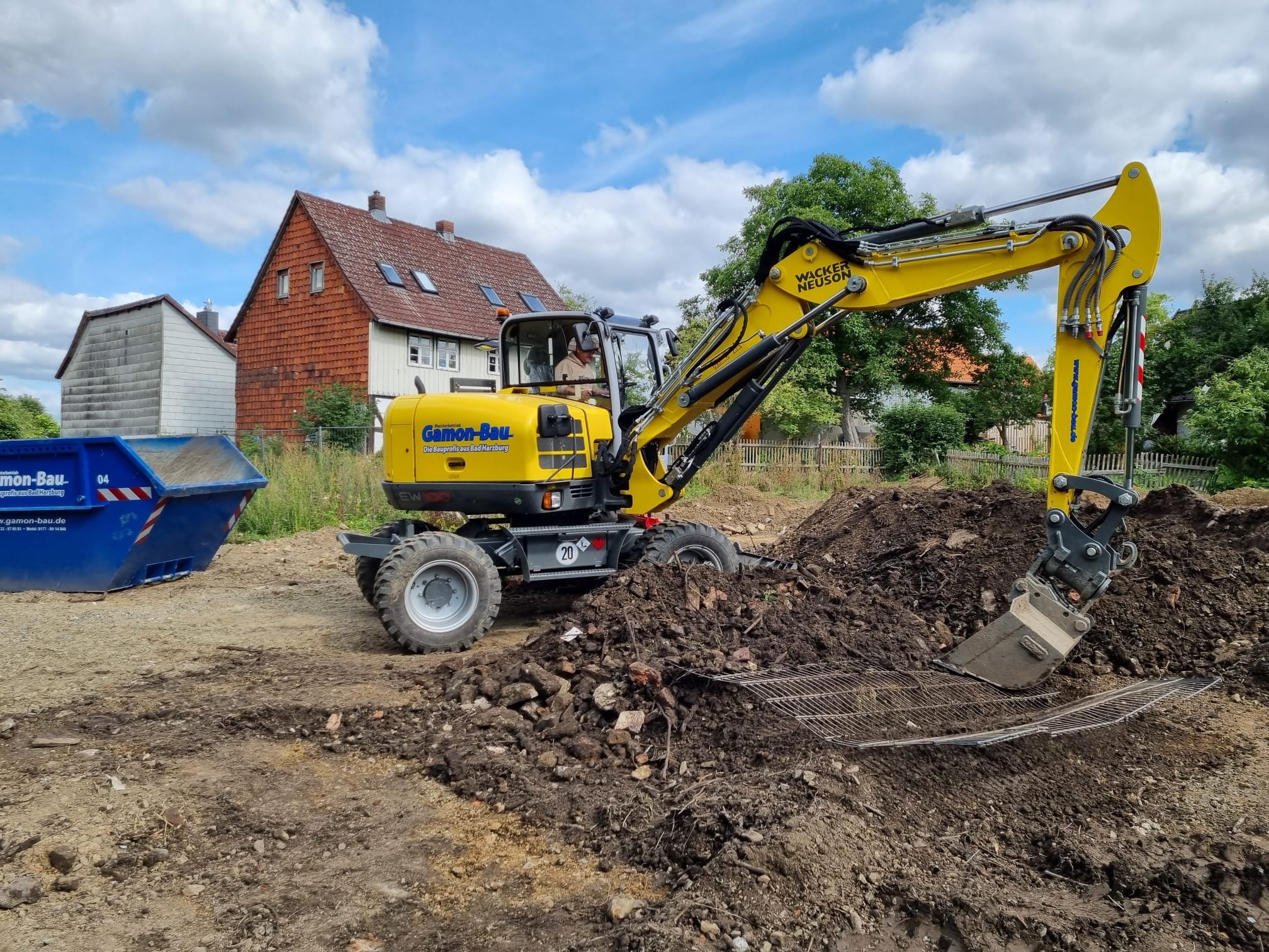 Bagger arbeitet auf einer Baustelle, schaufelt Erde in einen blauen Container, im Hintergrund sind Wohnhäuser und Bäume, Himmel ist bewölkt.